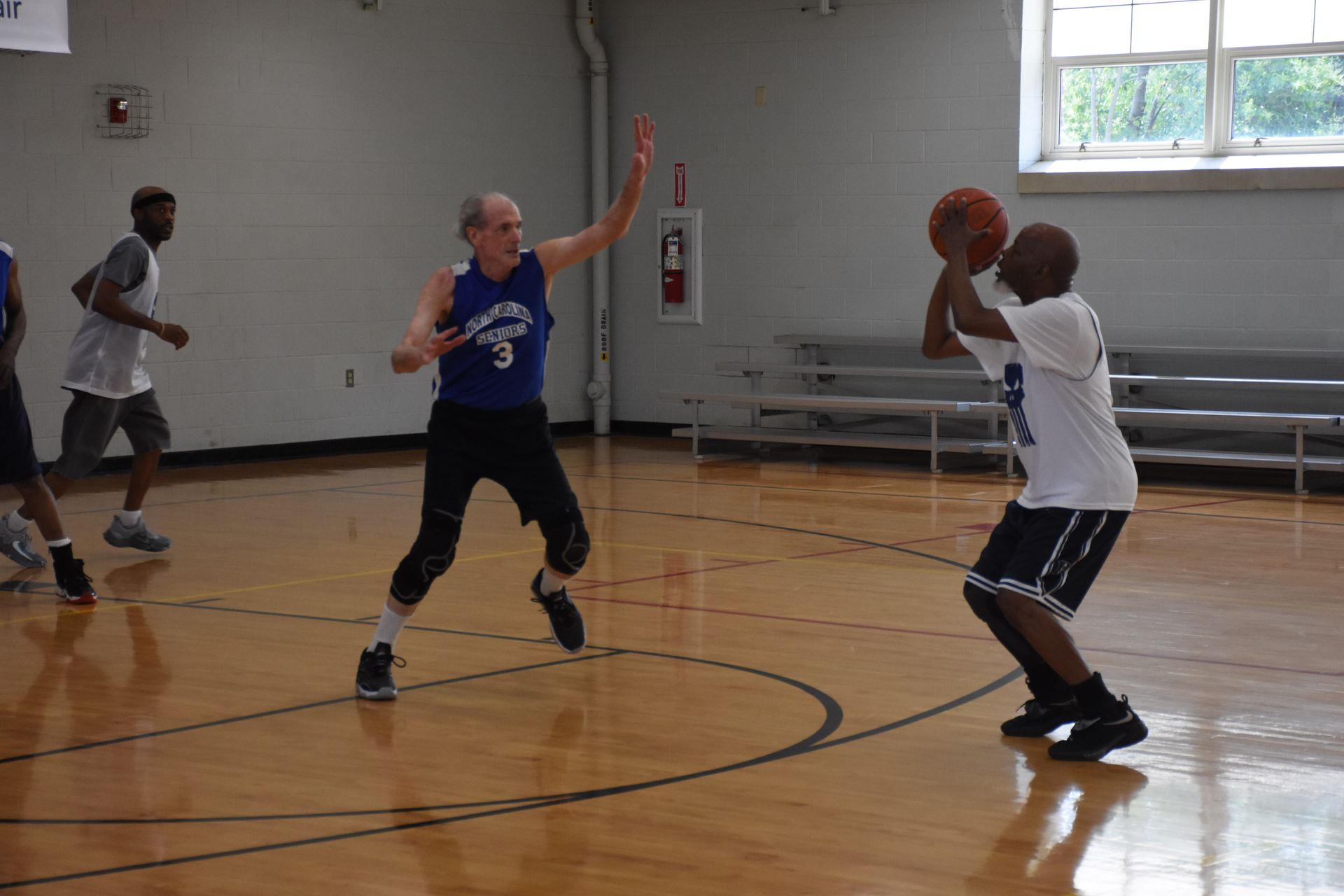 Basketball players on a wooden court. One shoots the ball, another defends, and a third runs.