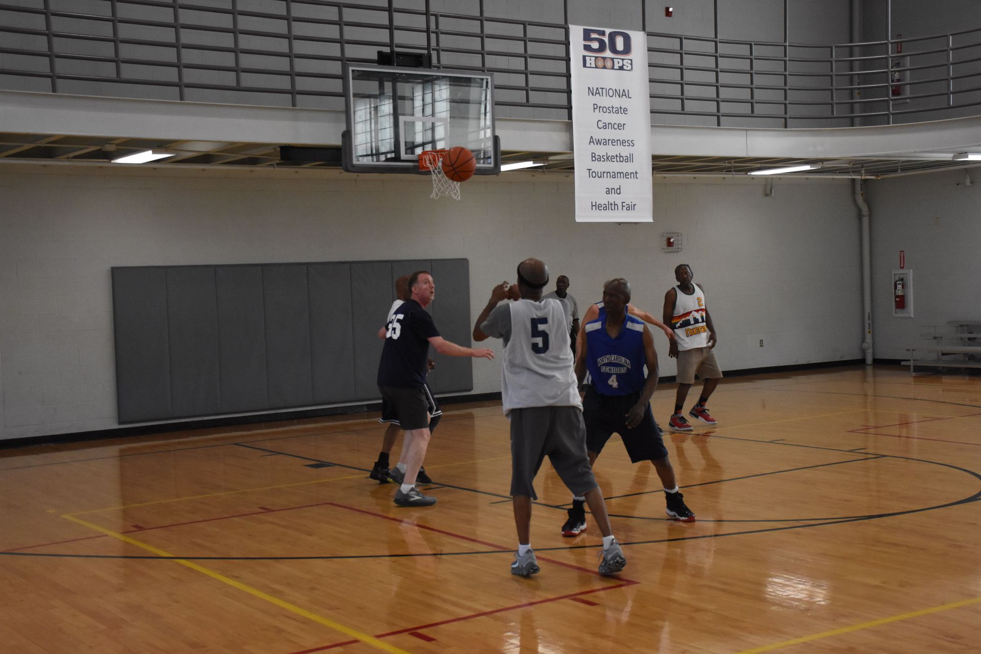 Basketball game in a gym: players, ball in air near hoop, hardwood floor, banner on wall.