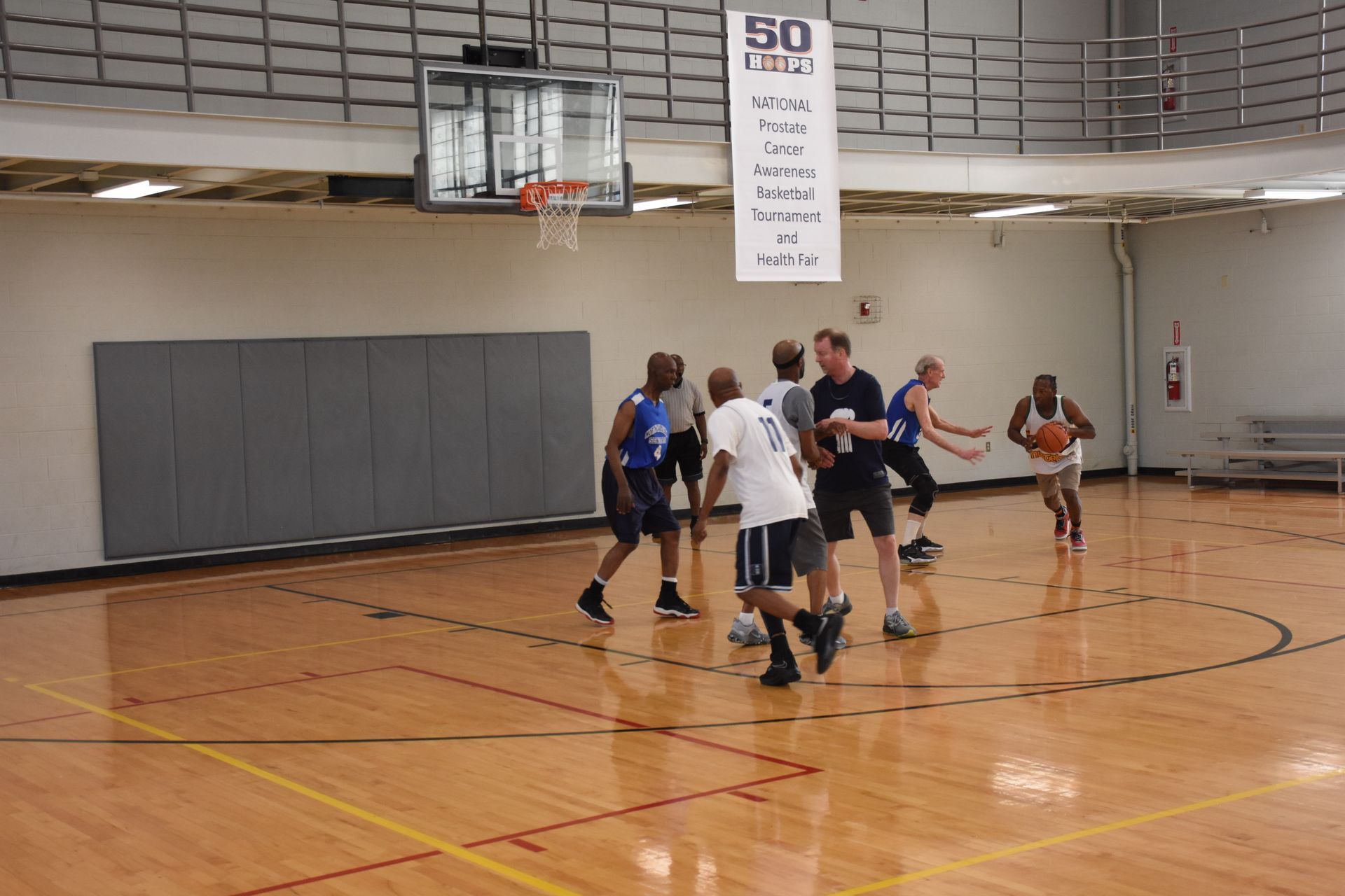Basketball game in progress: players on court, one dribbling, others near the basket, wooden floor, banner in the background.