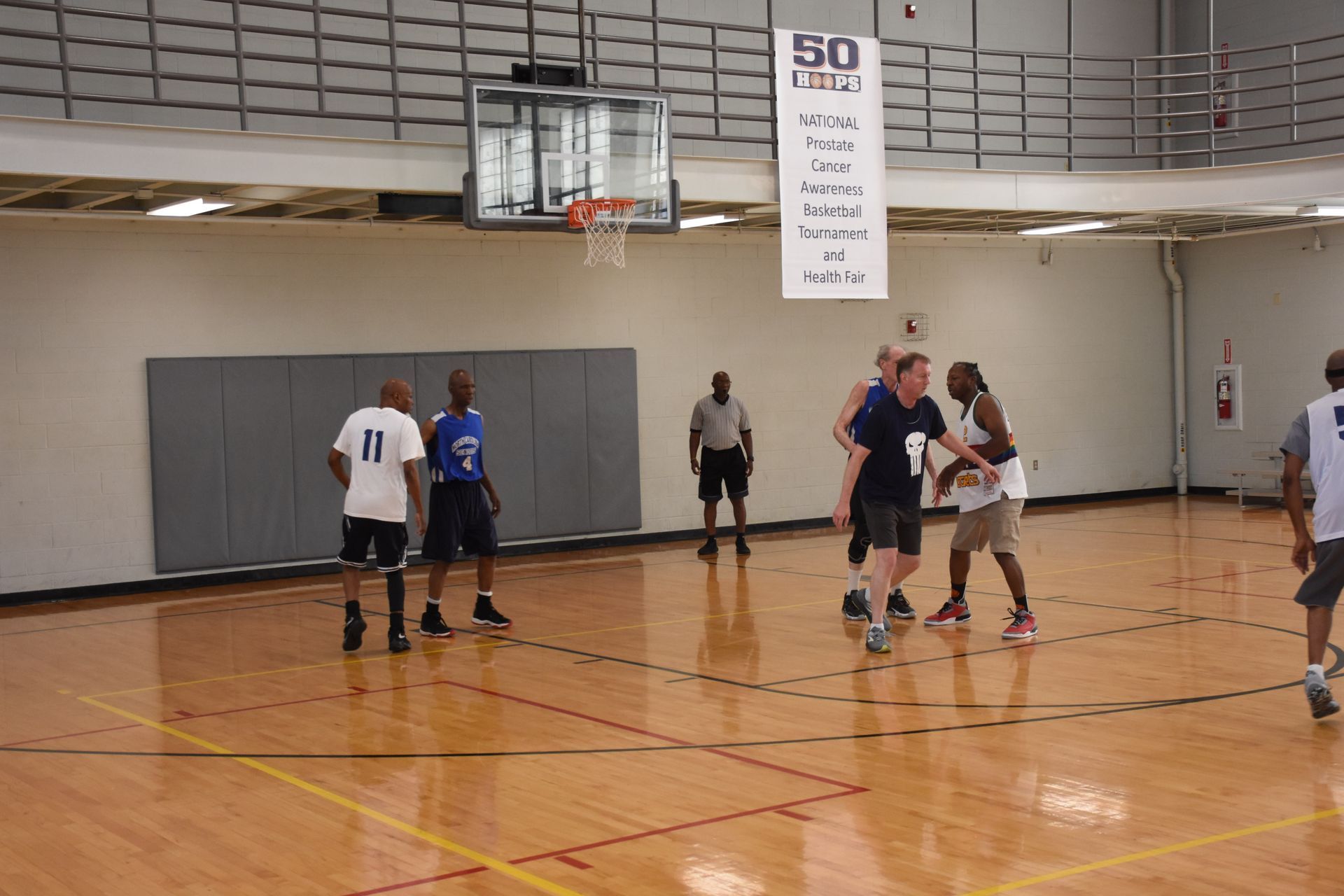 Basketball game in progress in an indoor court. Players in action, one with the ball near the center; a referee observes.