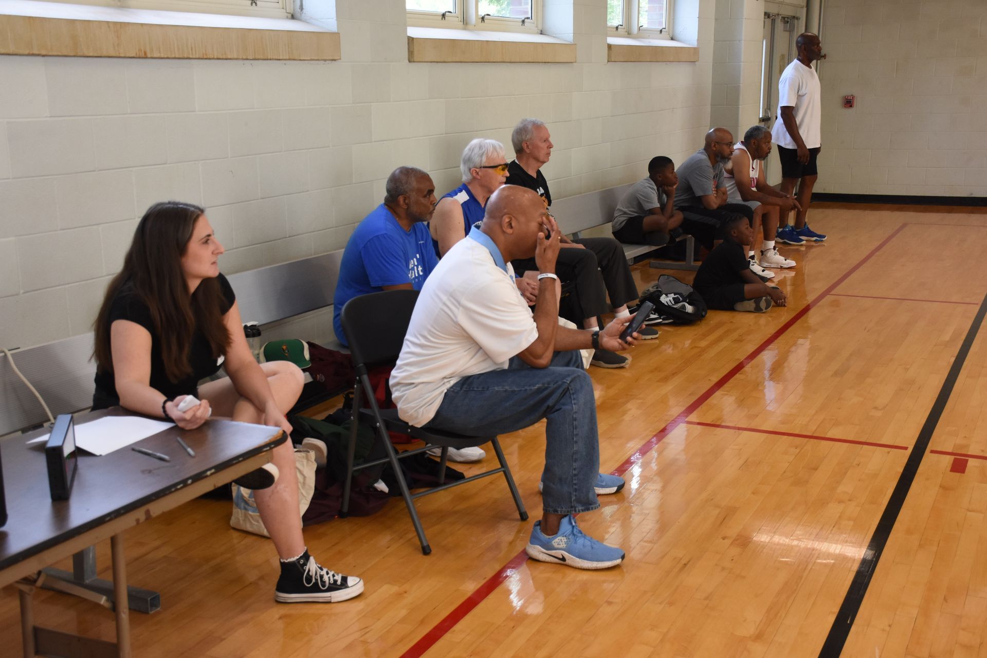 People seated on chairs, watching an event in a gymnasium, with a woman taking notes at a table.