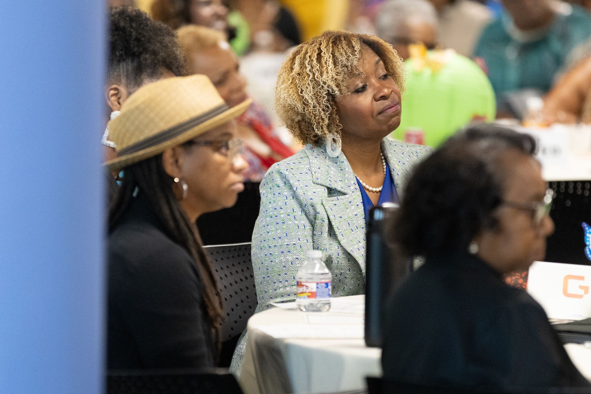 Attendees at a conference. Two women listen intently at a table. One in a fedora, another with blonde curly hair.