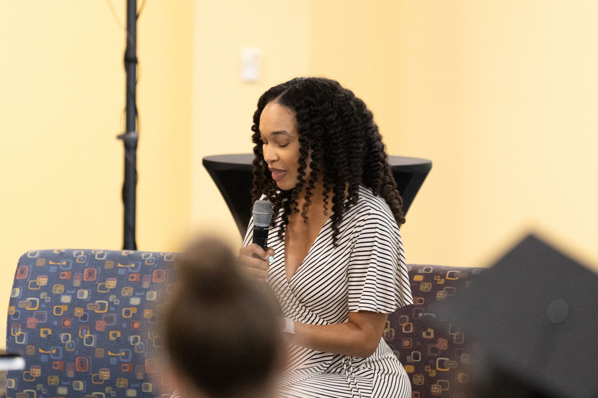 Woman speaking into a microphone, seated on a patterned sofa in a room with a light wall.