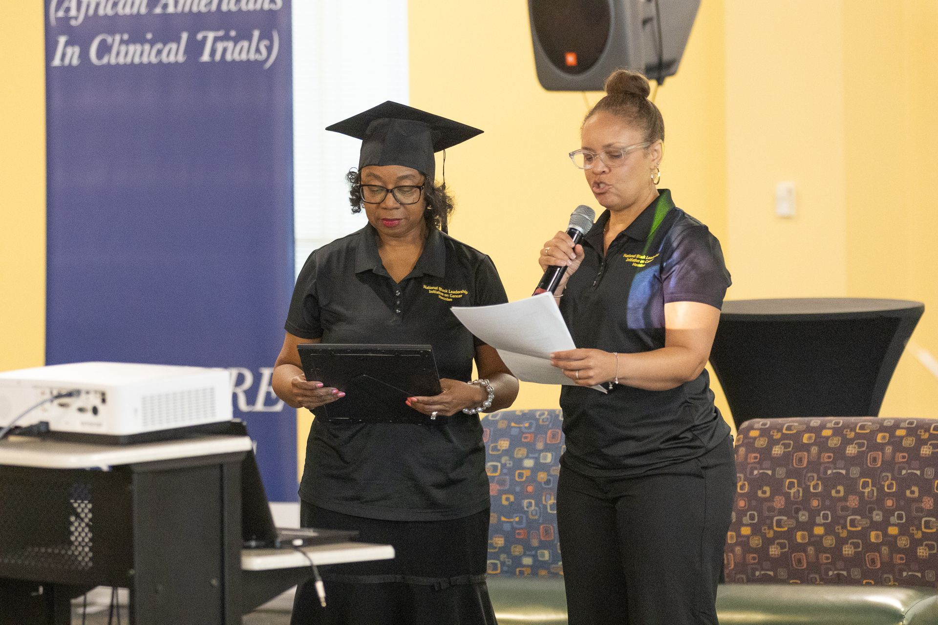 Two women at a presentation; one in a graduation cap. They're both holding papers. A projector and laptop are visible.