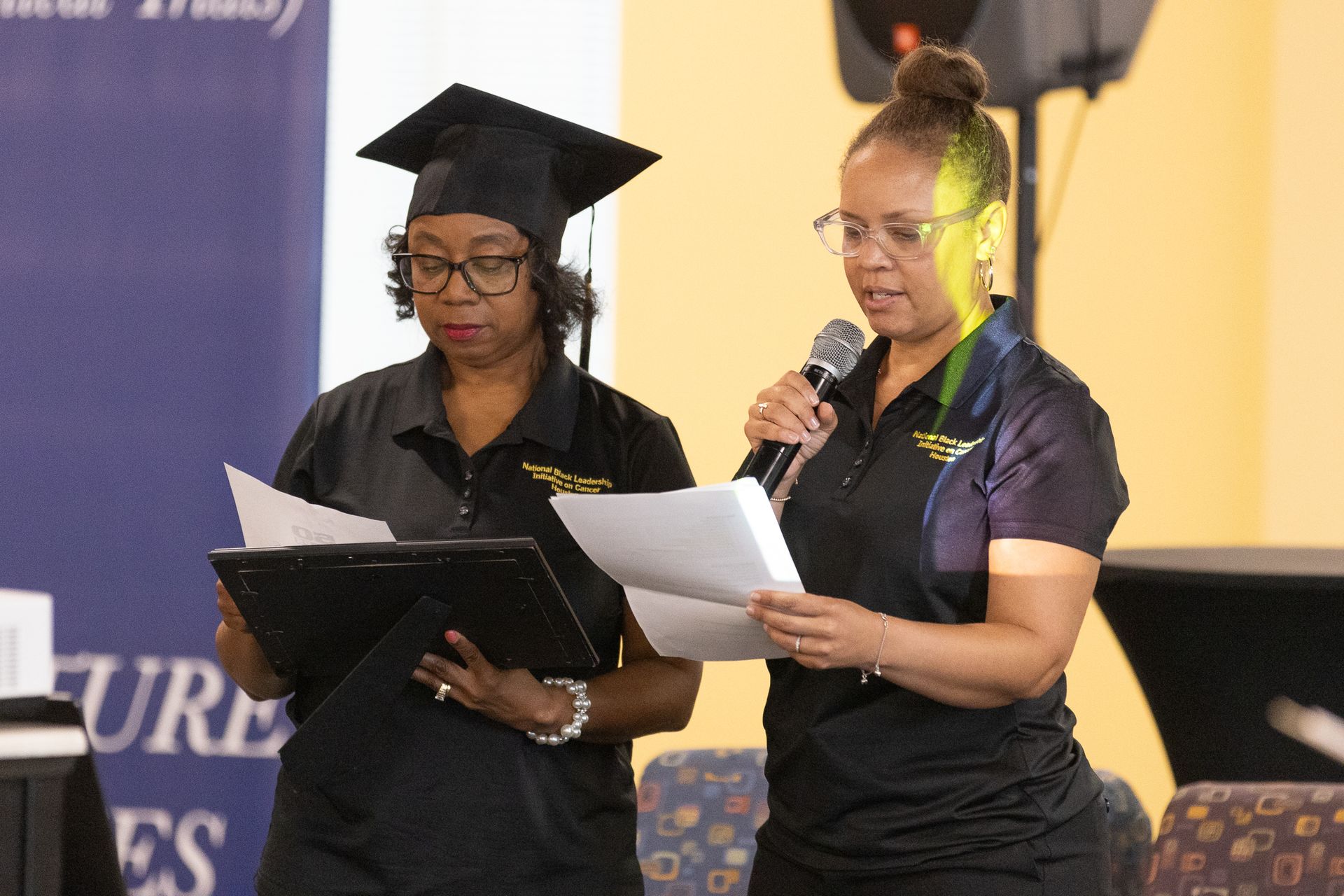 Two women, one in a graduation cap, reading from papers at an event.