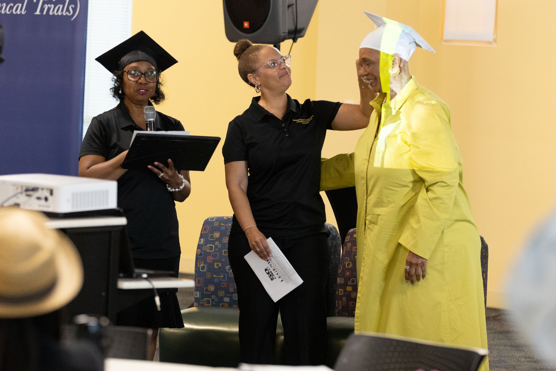 Woman in yellow robe has cap adjusted by another; woman speaks at podium.