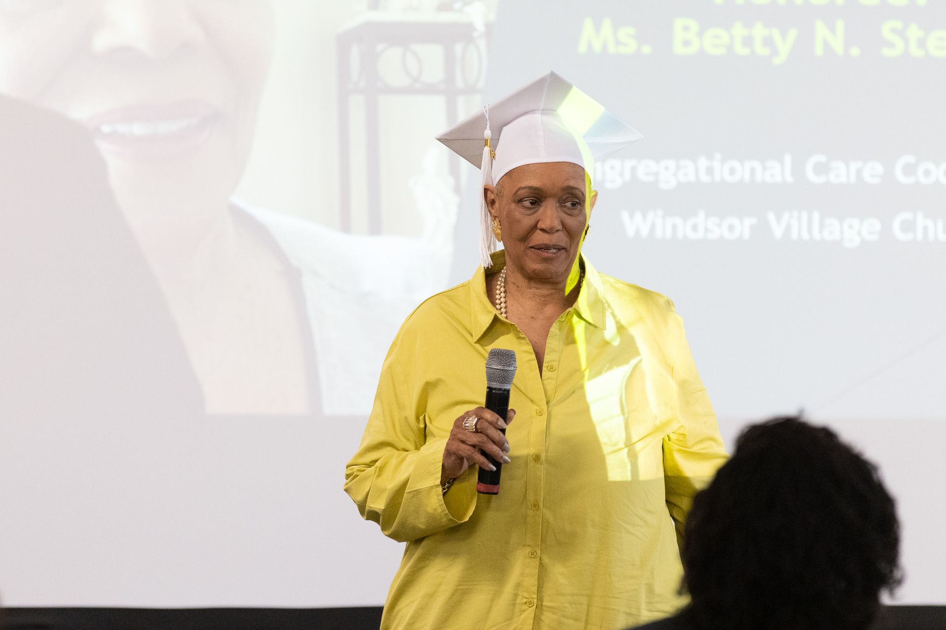 Woman in yellow shirt and graduation cap speaking into a microphone.