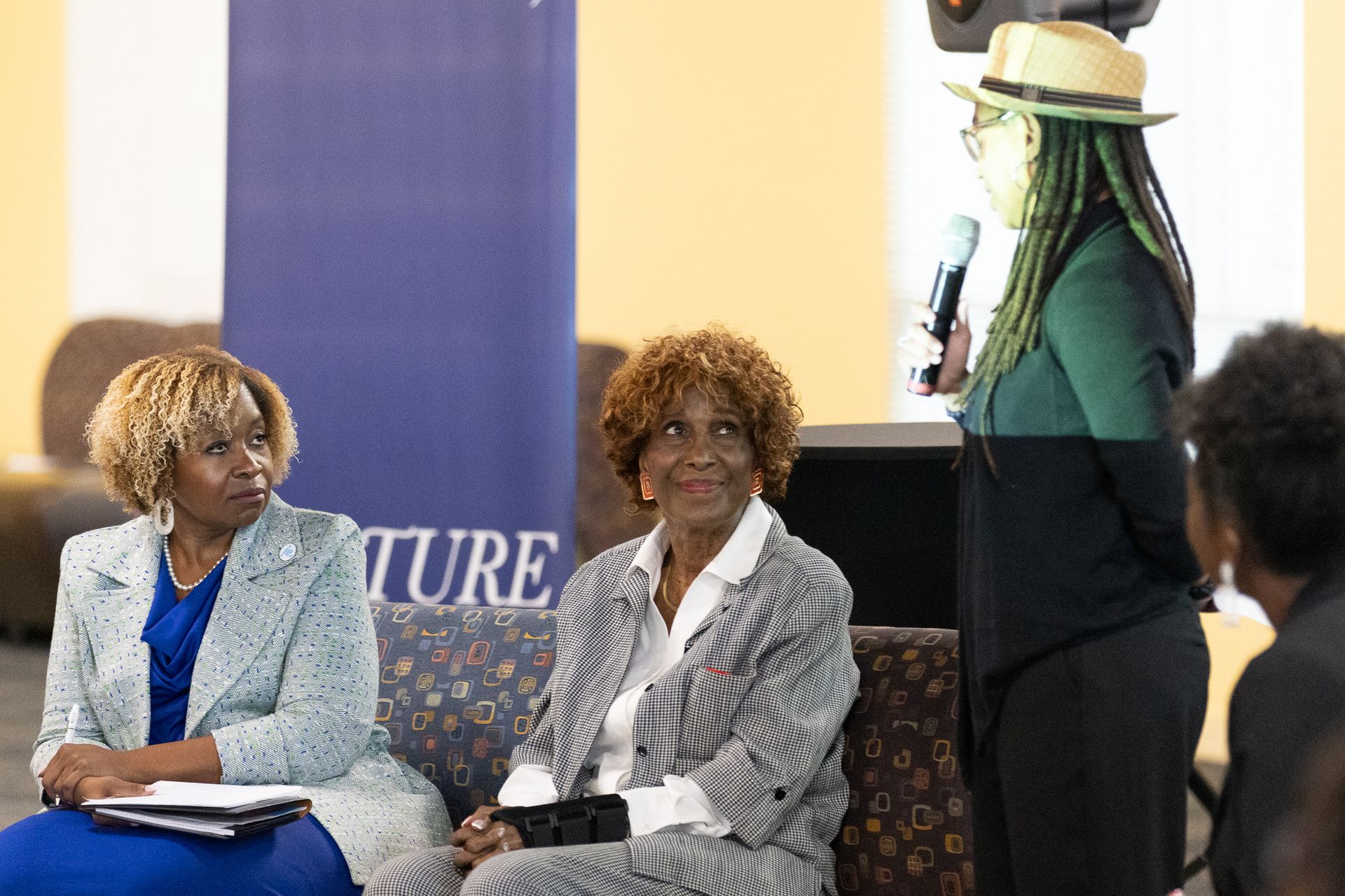 Three women at an event: two seated, listening, and one standing, speaking into a microphone.