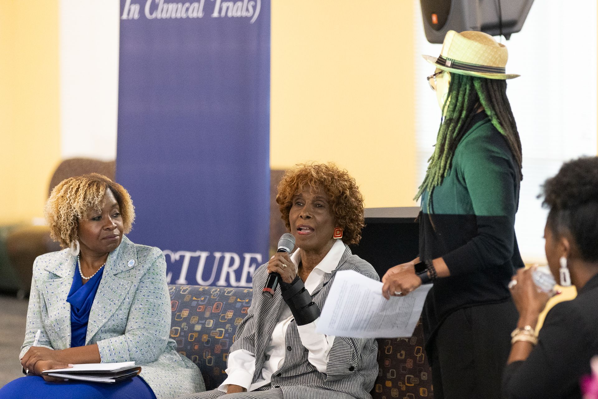 Panel of women at a conference. One speaks into a mic, another writes. A fourth woman stands nearby.