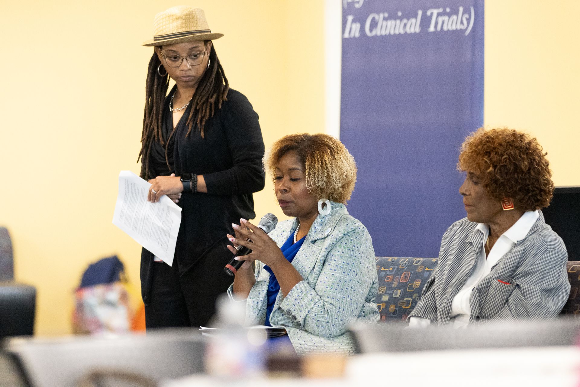 Three women participate in a discussion at a table; one speaks into a microphone. Another woman stands and holds papers.
