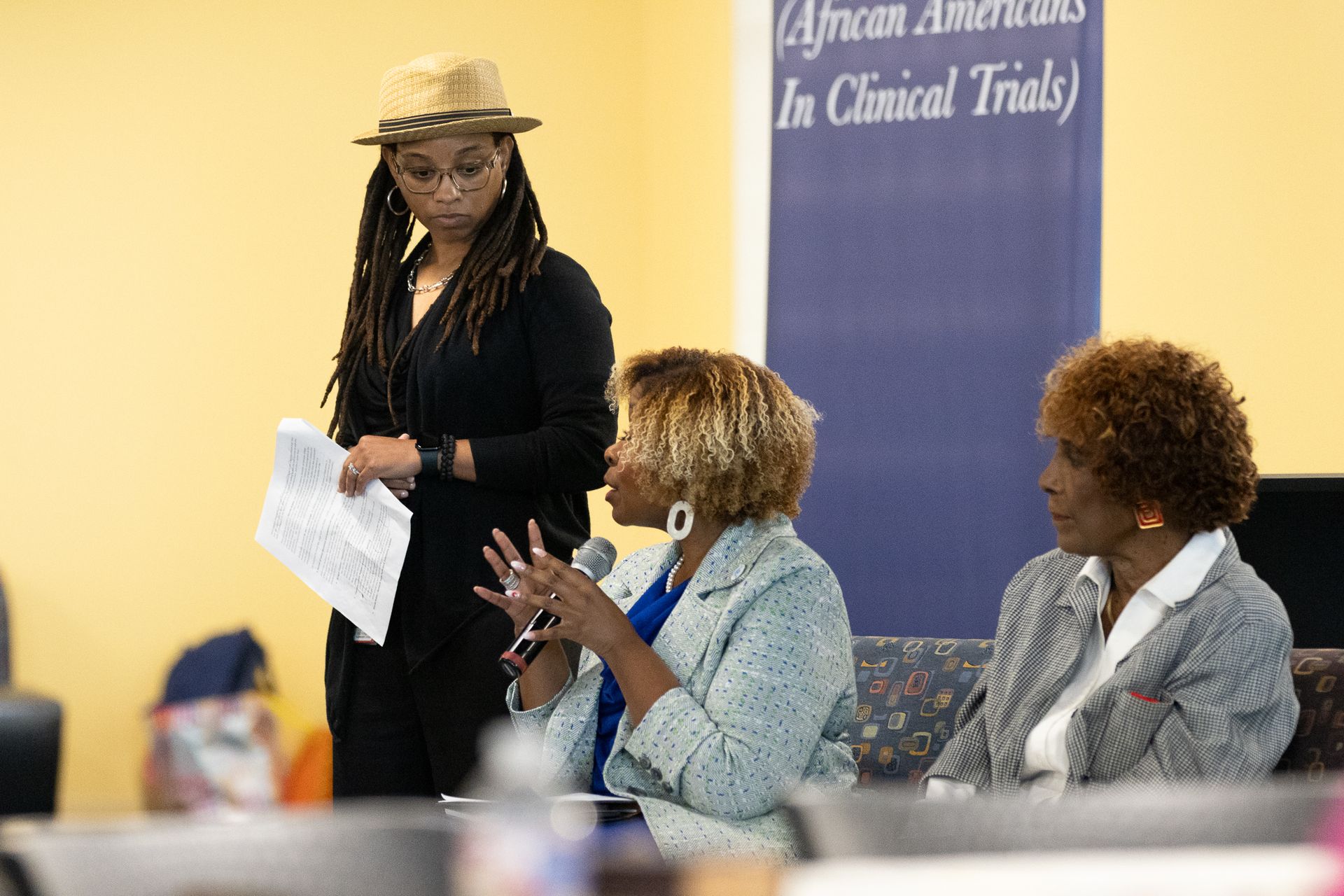 Panel discussion: Three women seated, one speaking. Woman standing at lectern. Banner in background.