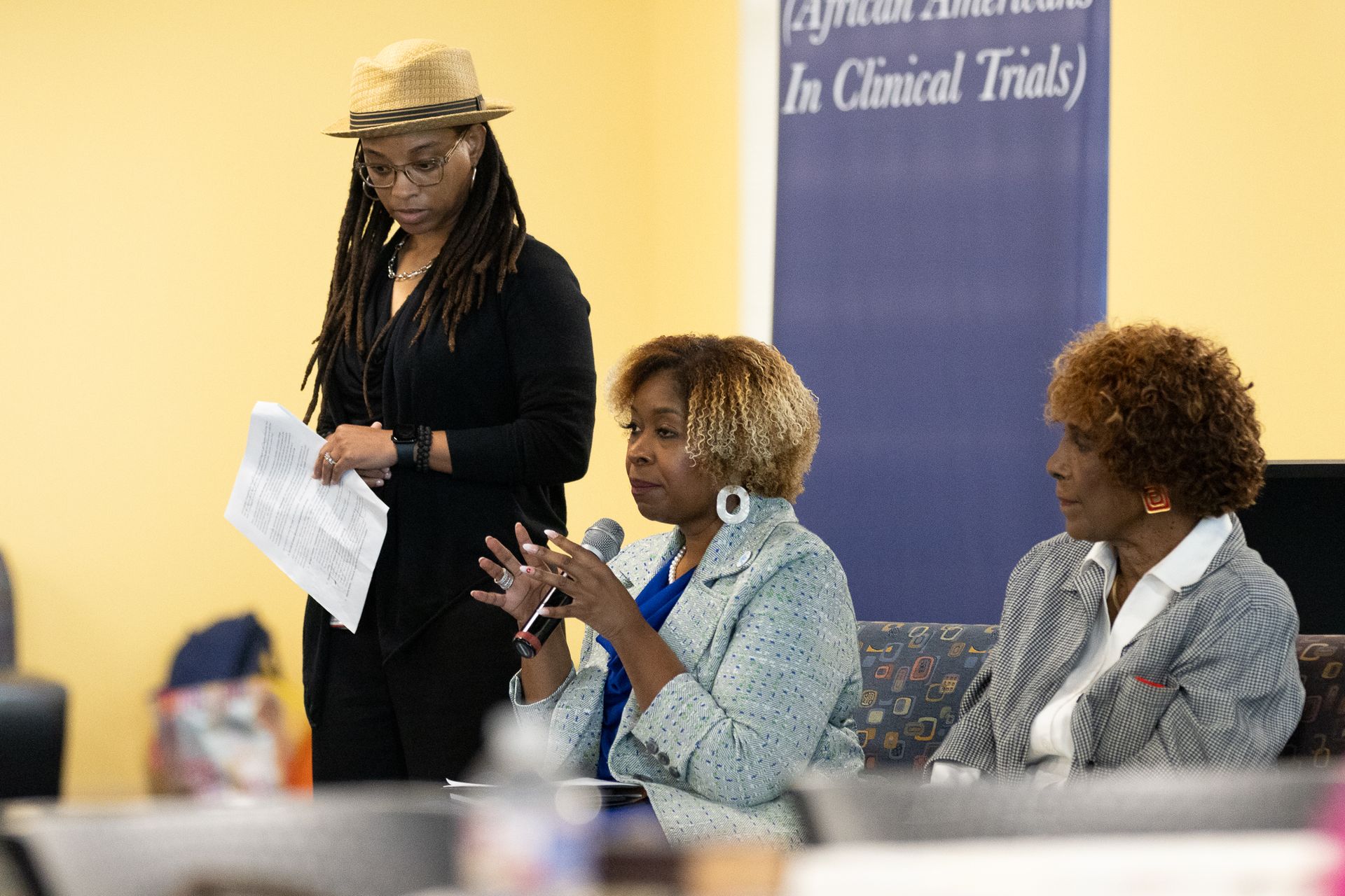 Three women at a panel discussion, one speaking into a mic. A banner behind them.