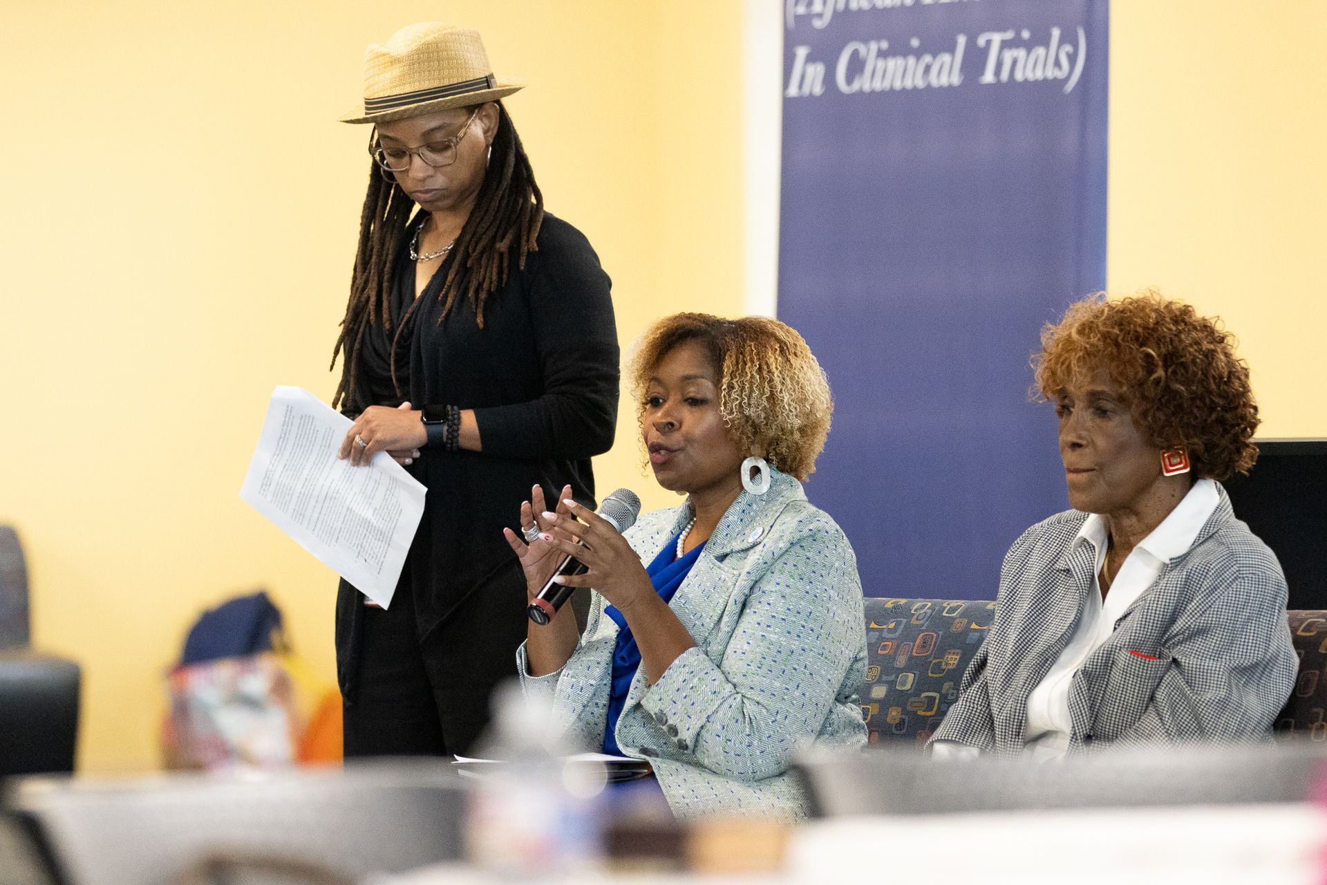 Three women at a panel discussion; one speaks into a microphone, another listens, the third stands behind them.