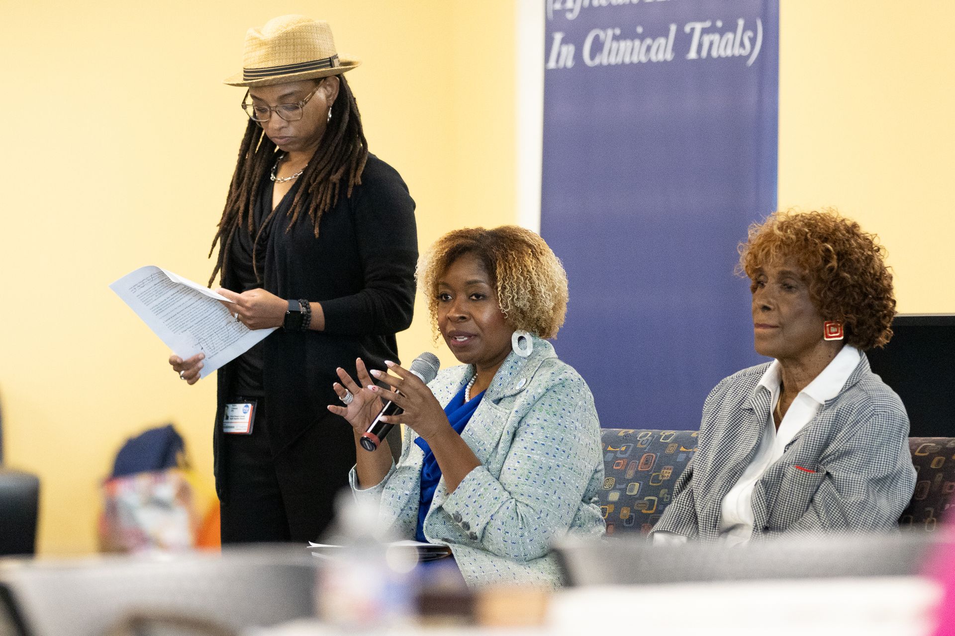 Three women at a panel discussion, one speaking into a microphone. Banner in background: 