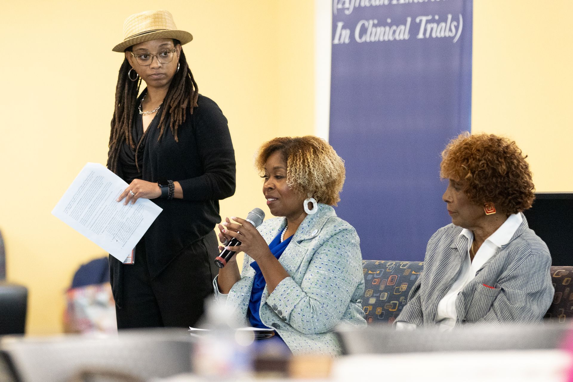 Three people at a table; one speaks into a mic, another observes, and one stands with papers. Background banner.