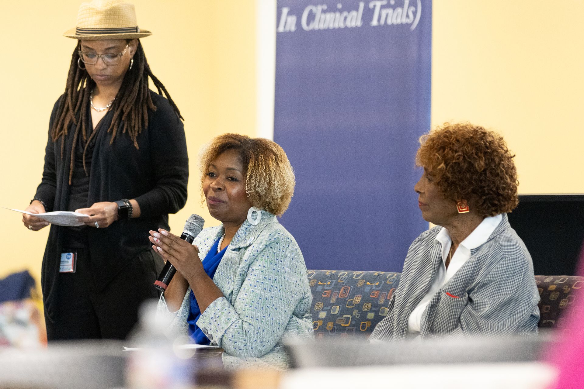 Three women at a panel discussion; one speaking into a microphone, another listening, and a third standing.