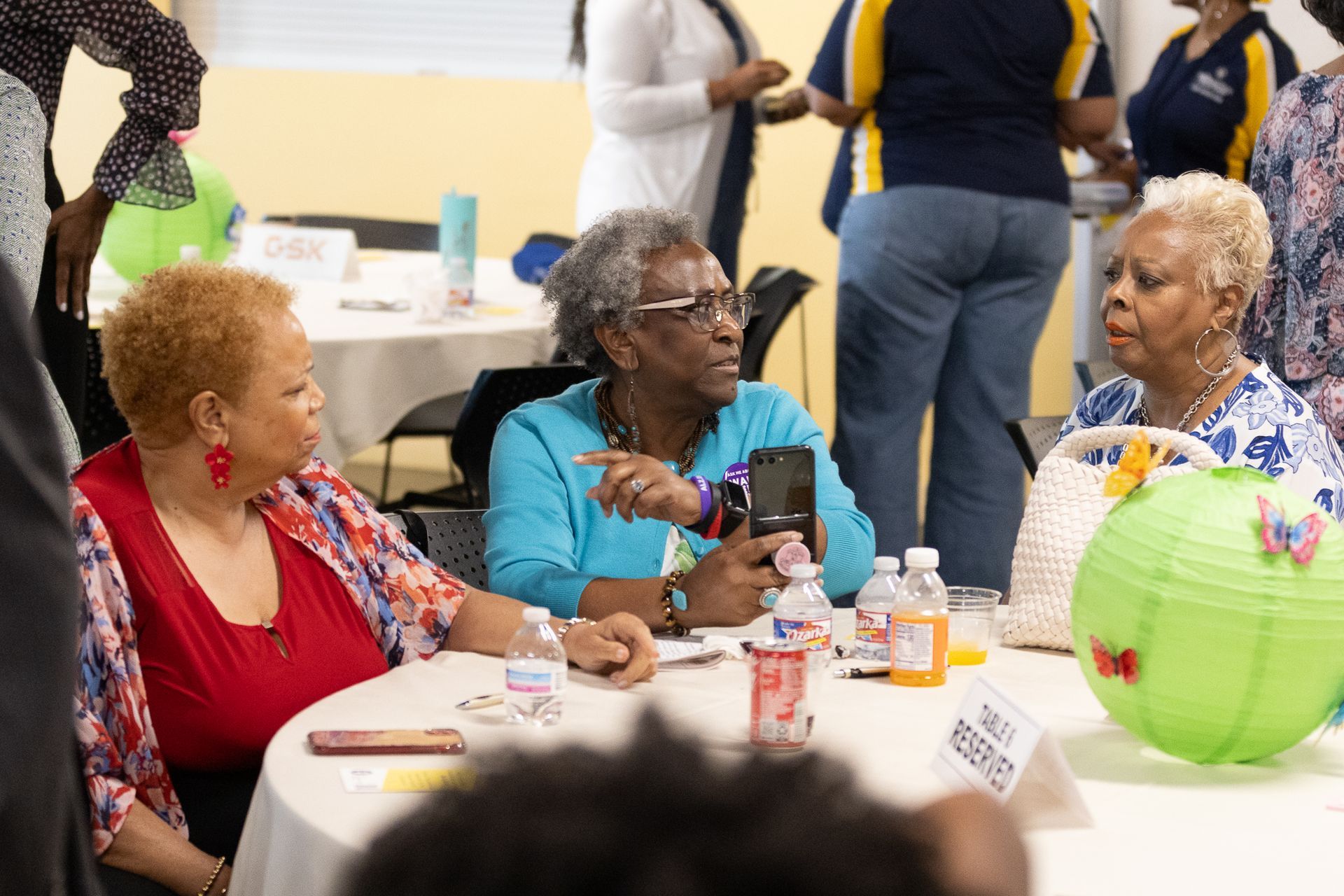 Three women seated at a table, conversing. One holds a phone. Setting is a social gathering with decorations.