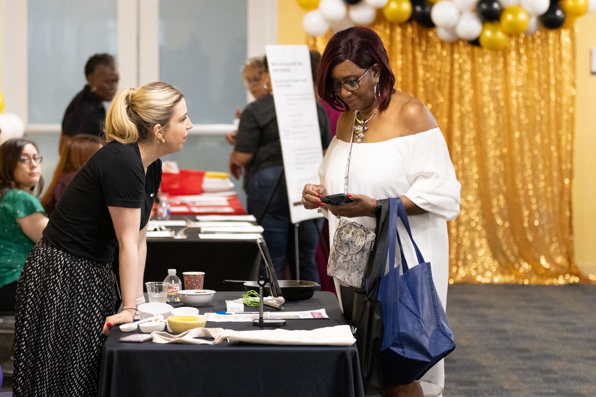 Woman in white dress looks at phone while another woman speaks at an event table with gold backdrop.