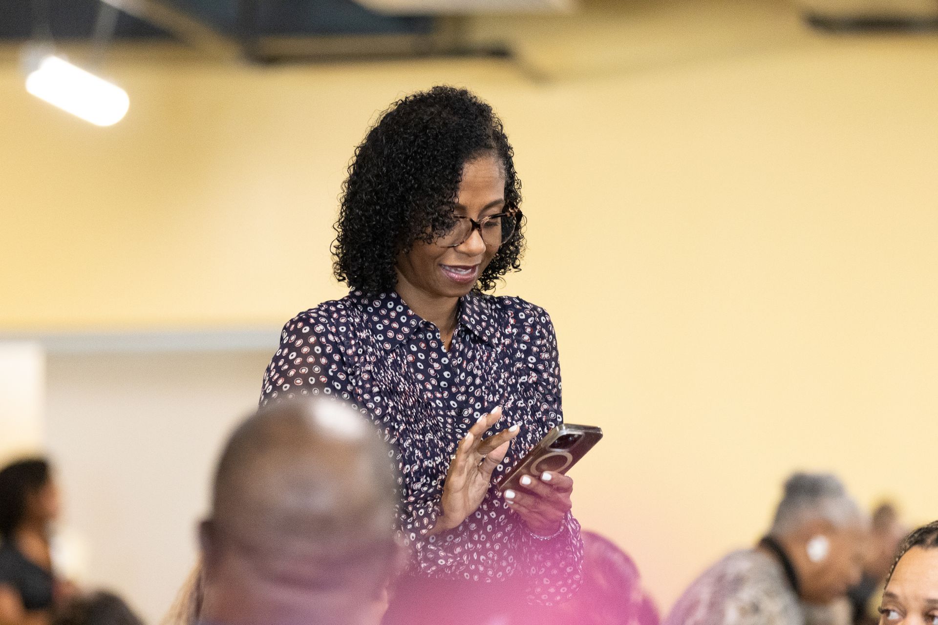 Woman in glasses, patterned shirt, using phone, in a room with other people.