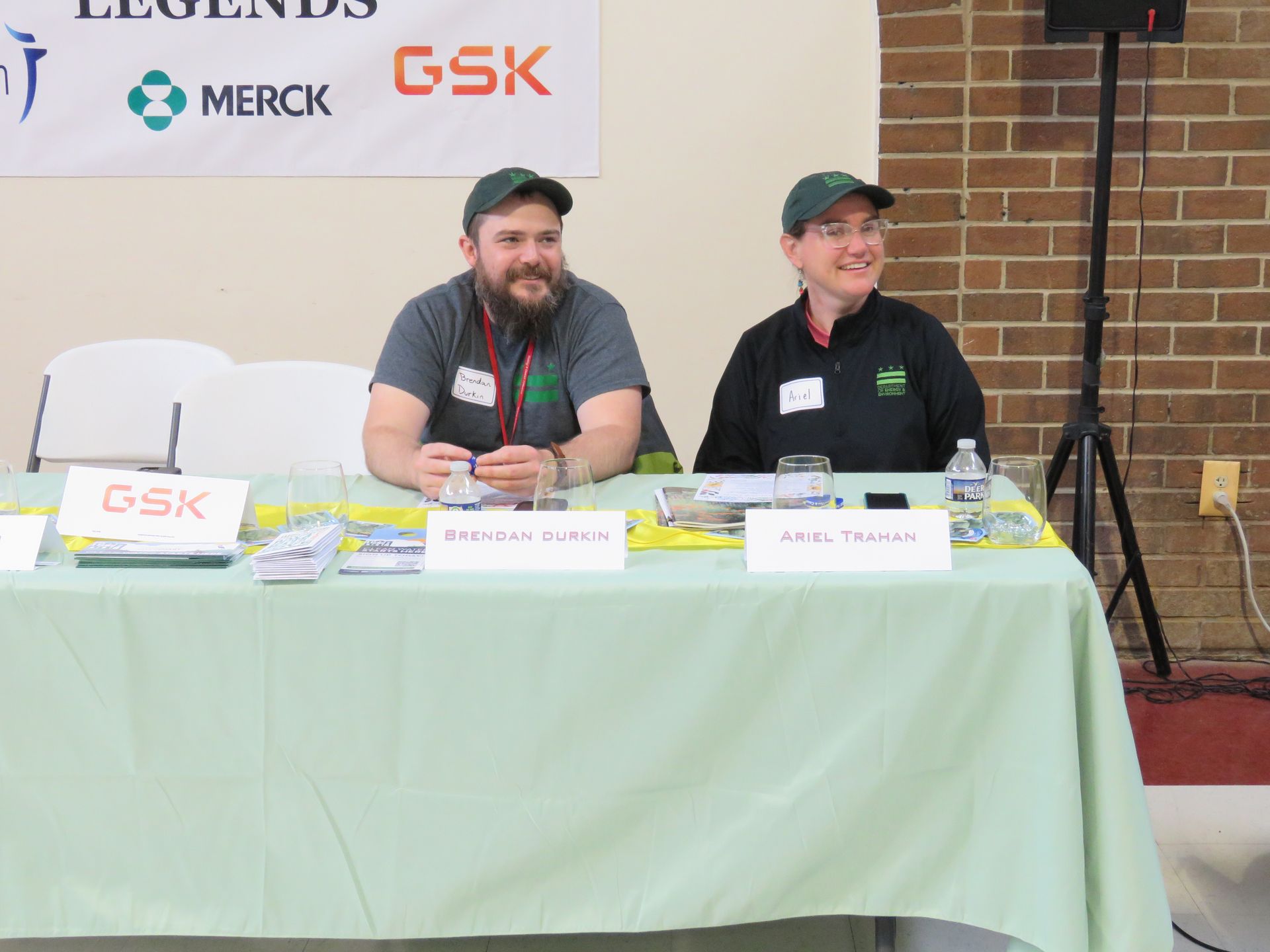 Two people at a table; one with a beard, both wearing green shirts, under a banner for GSK and Merck.