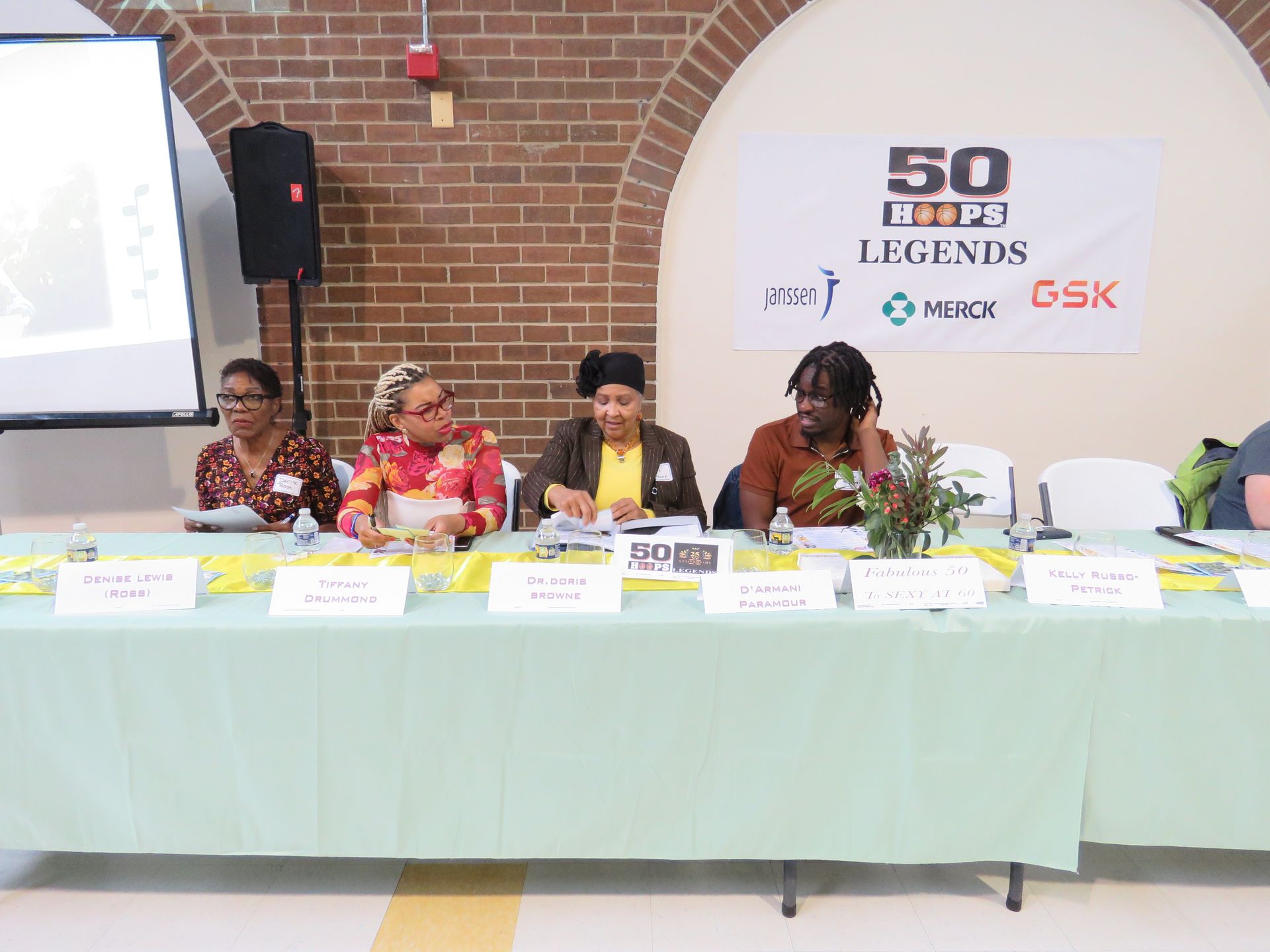 Panel of four people seated at a table with name placards; banner reads 