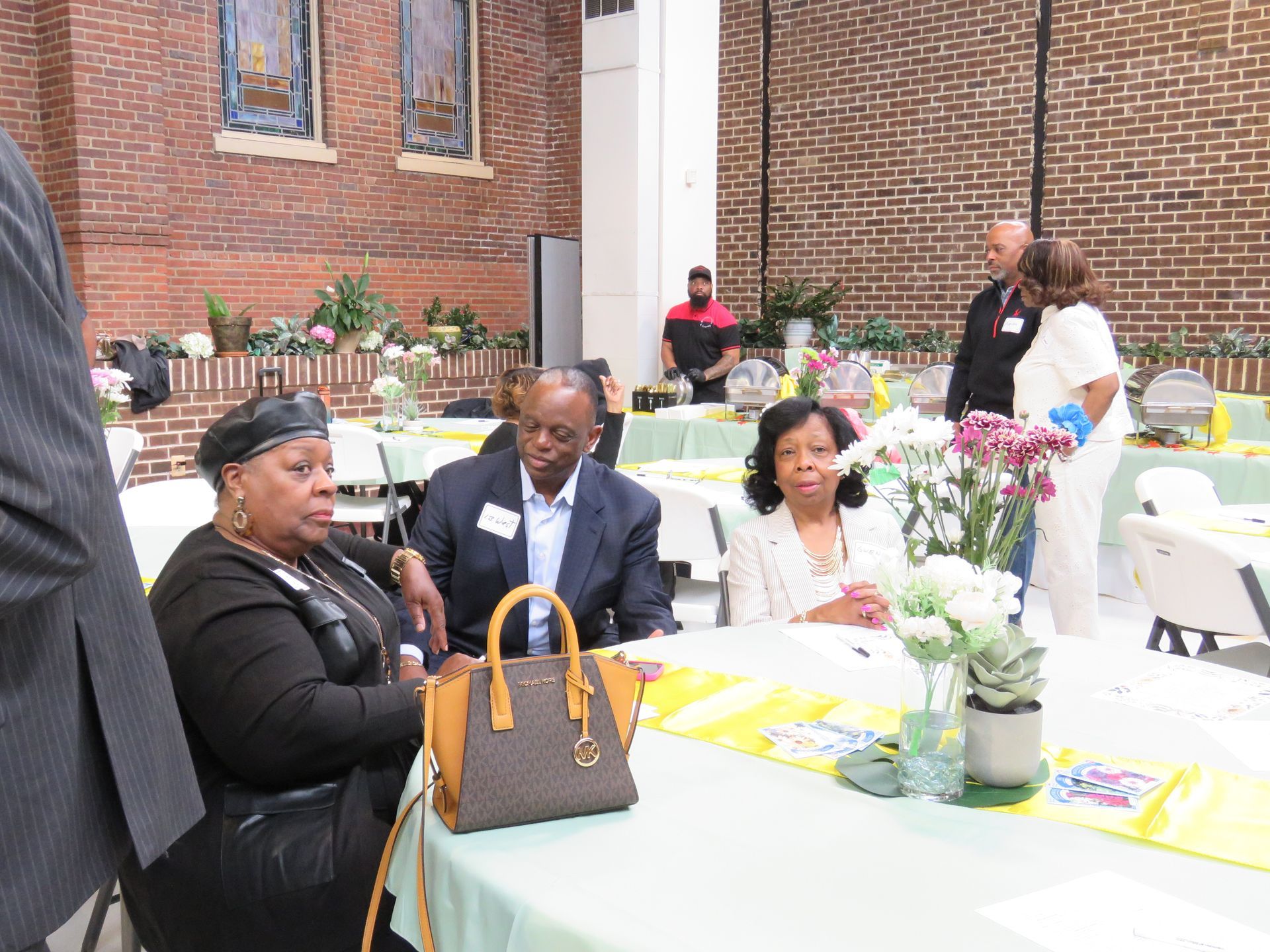 People seated at decorated tables in a brick-walled room. Flowers, food, and staff visible.