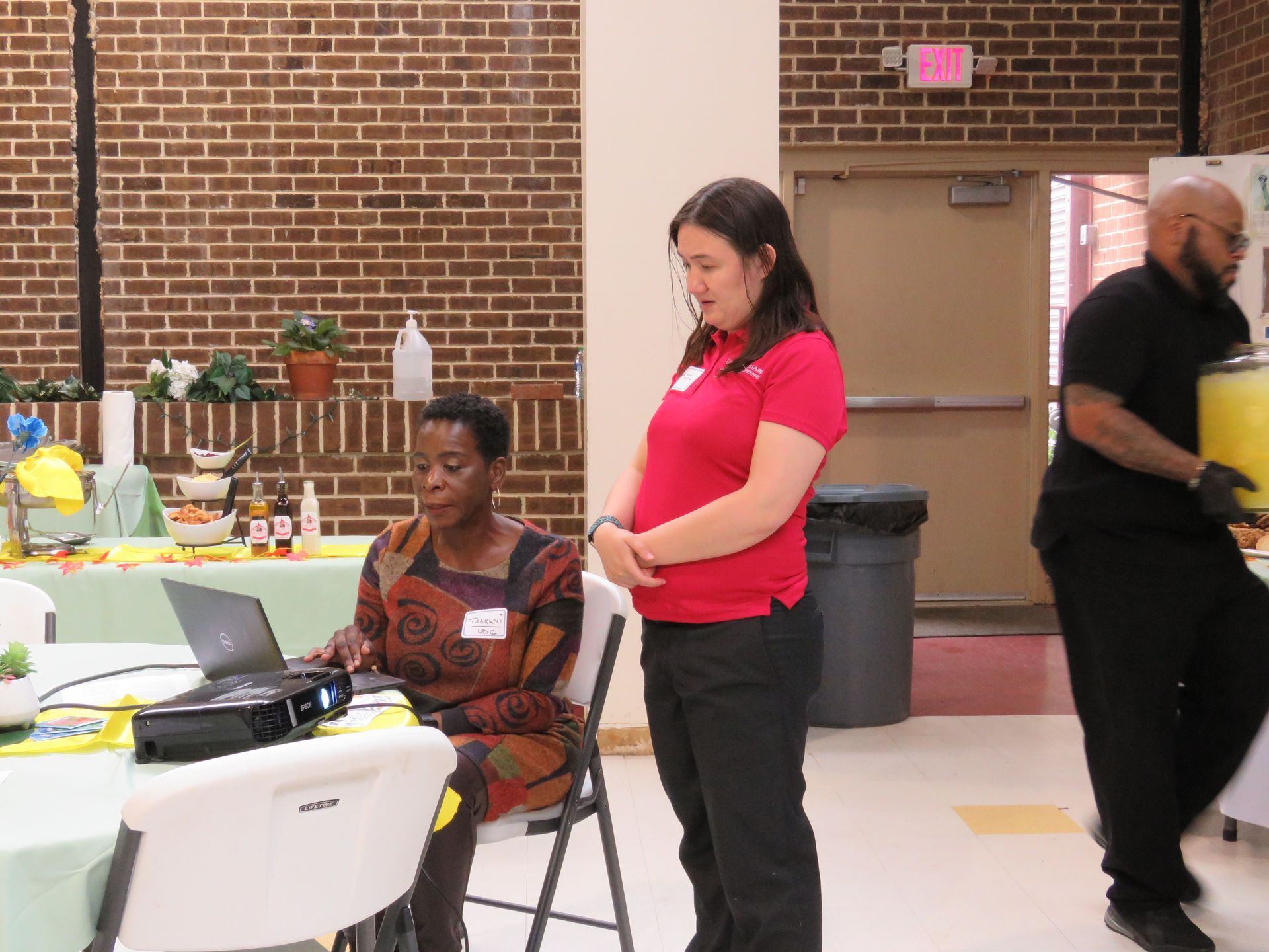 Woman using laptop, two others stand nearby, event setup in a brick-walled room.