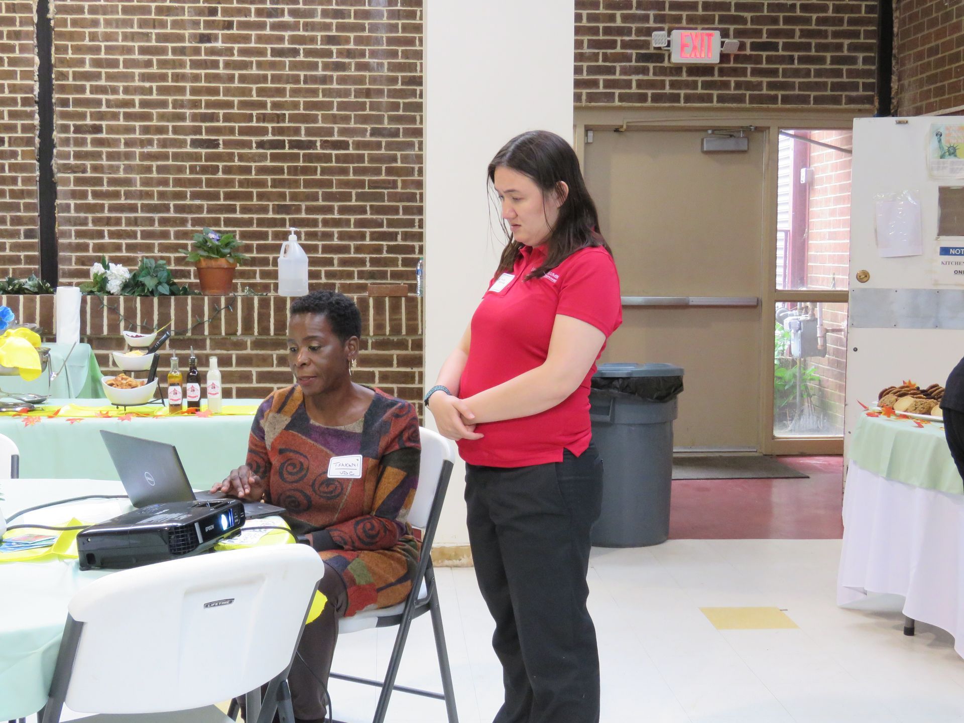 Woman using laptop, another woman watches in a room with a buffet table and an exit door.
