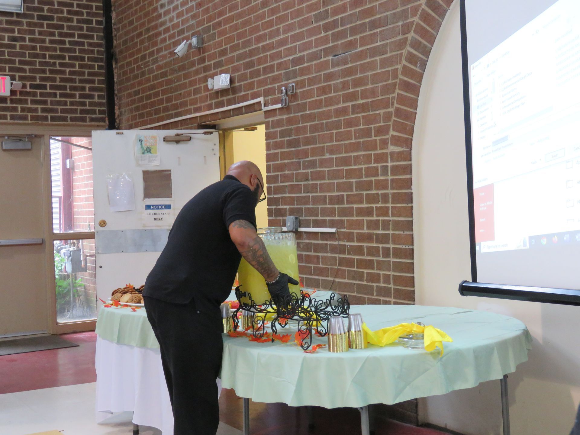 Man setting up at a table with a green tablecloth in front of a brick wall and projector screen.