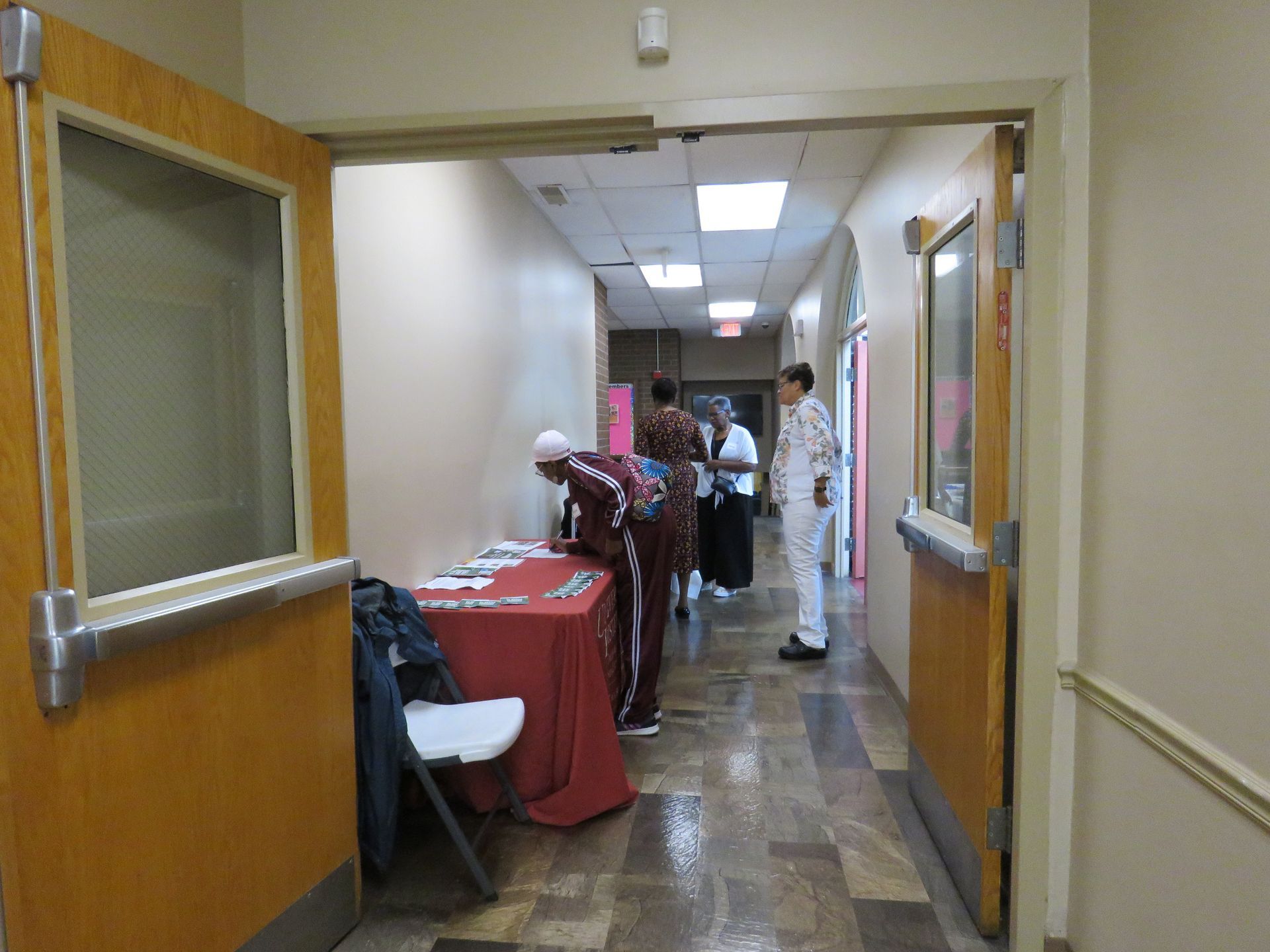 Hallway with people near a table, doors on each side, linoleum floor.