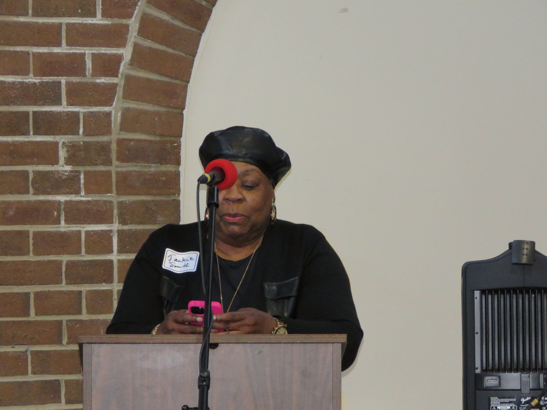 Woman speaking at a lectern, holding phone. Brick wall and arch in background. Black outfit, microphone.