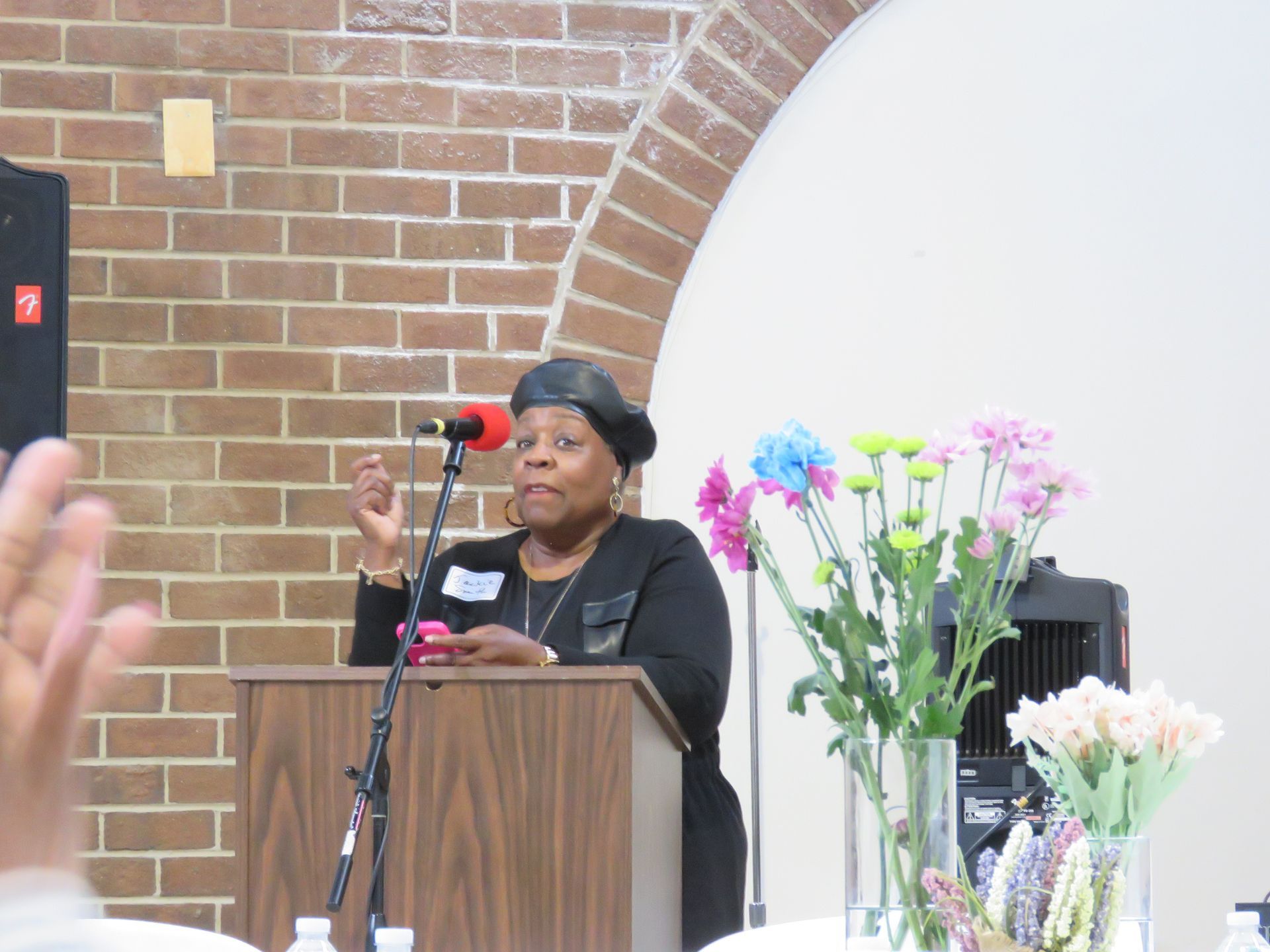 Woman speaking at a podium, brick wall background, flowers, and a person's raised hand.