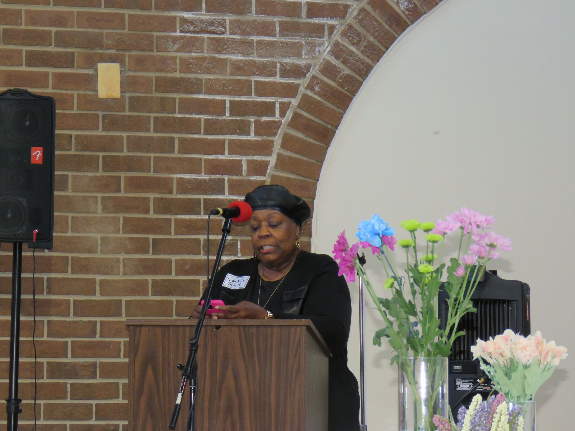 Woman speaking at a podium with microphone in front of brick wall, flowers in a vase to her right.