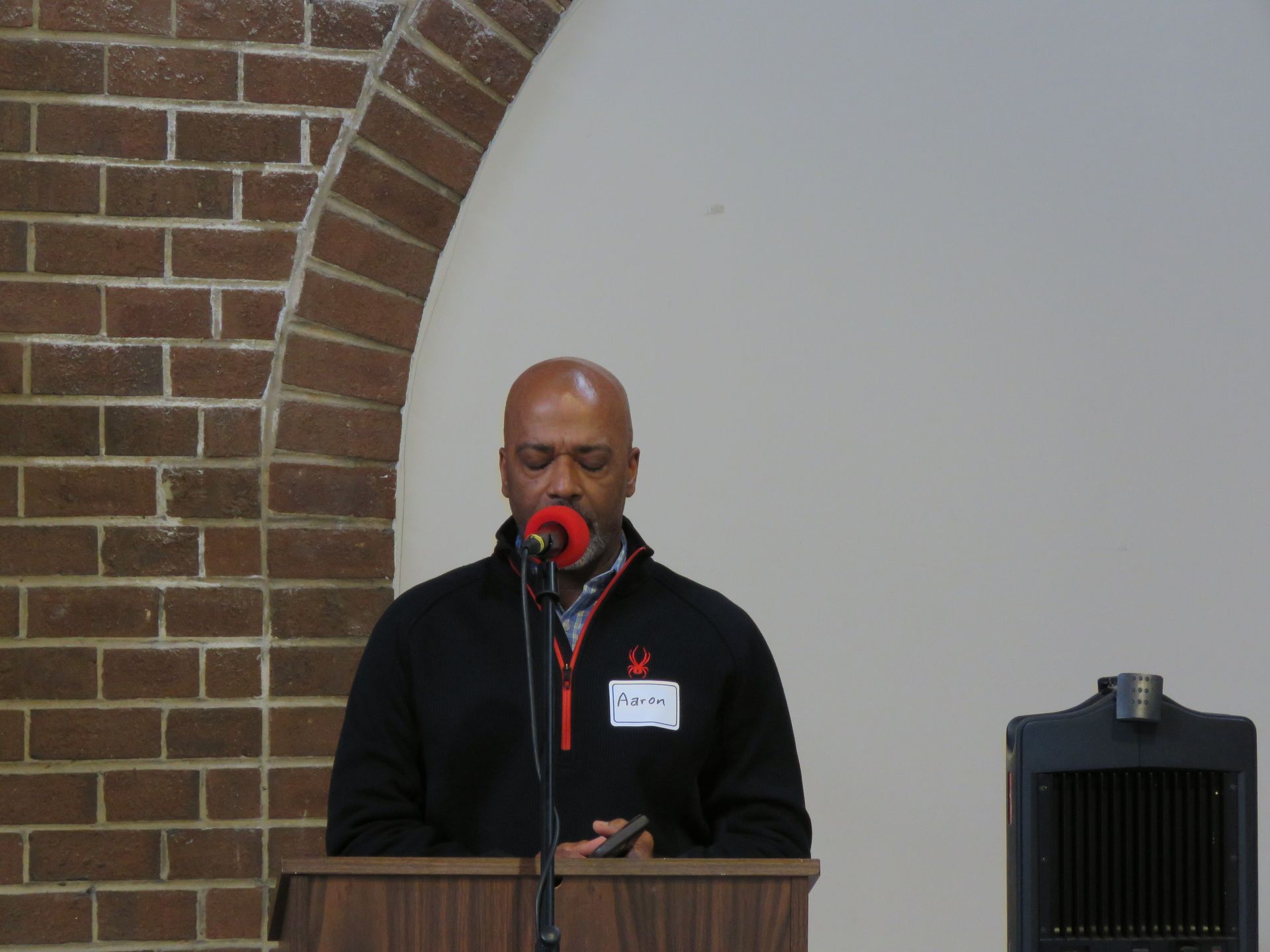 Man speaking at a podium; brick archway background, black sweater, name tag visible.