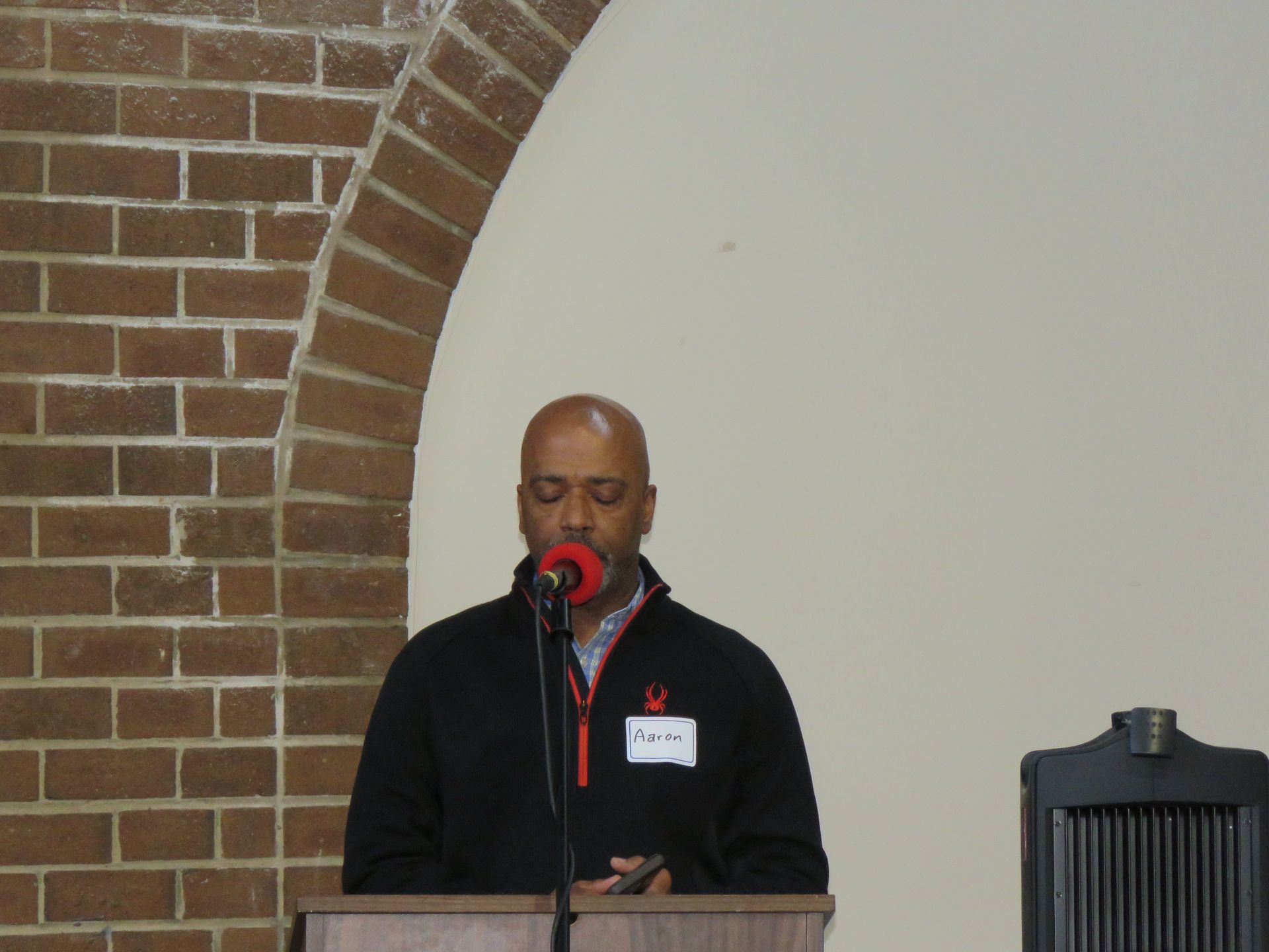 Man speaking at a podium with a red ribbon pin, brick and arched wall background.