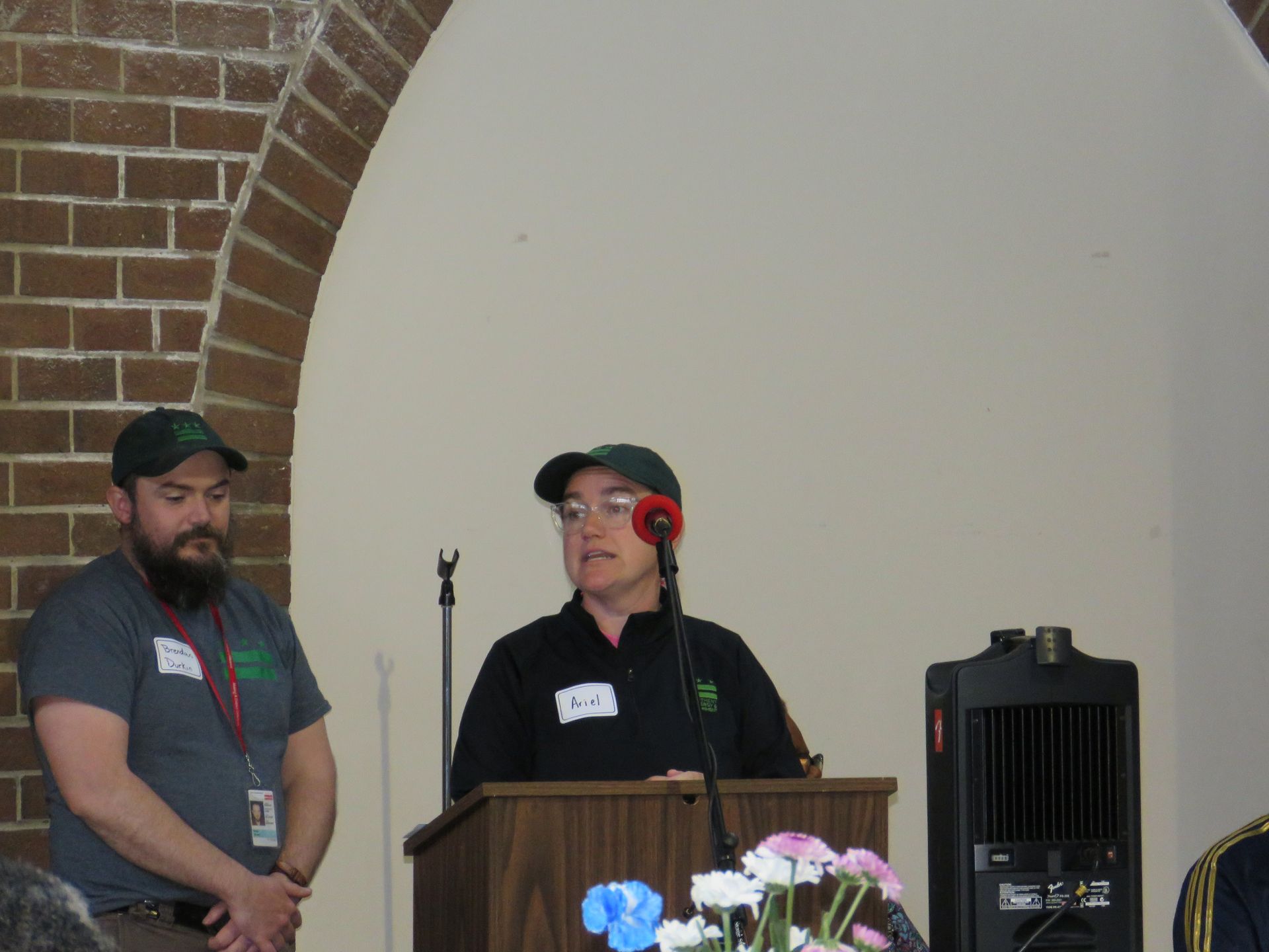 Two people wearing green hats at a podium, speaking into a microphone in front of a brick archway.
