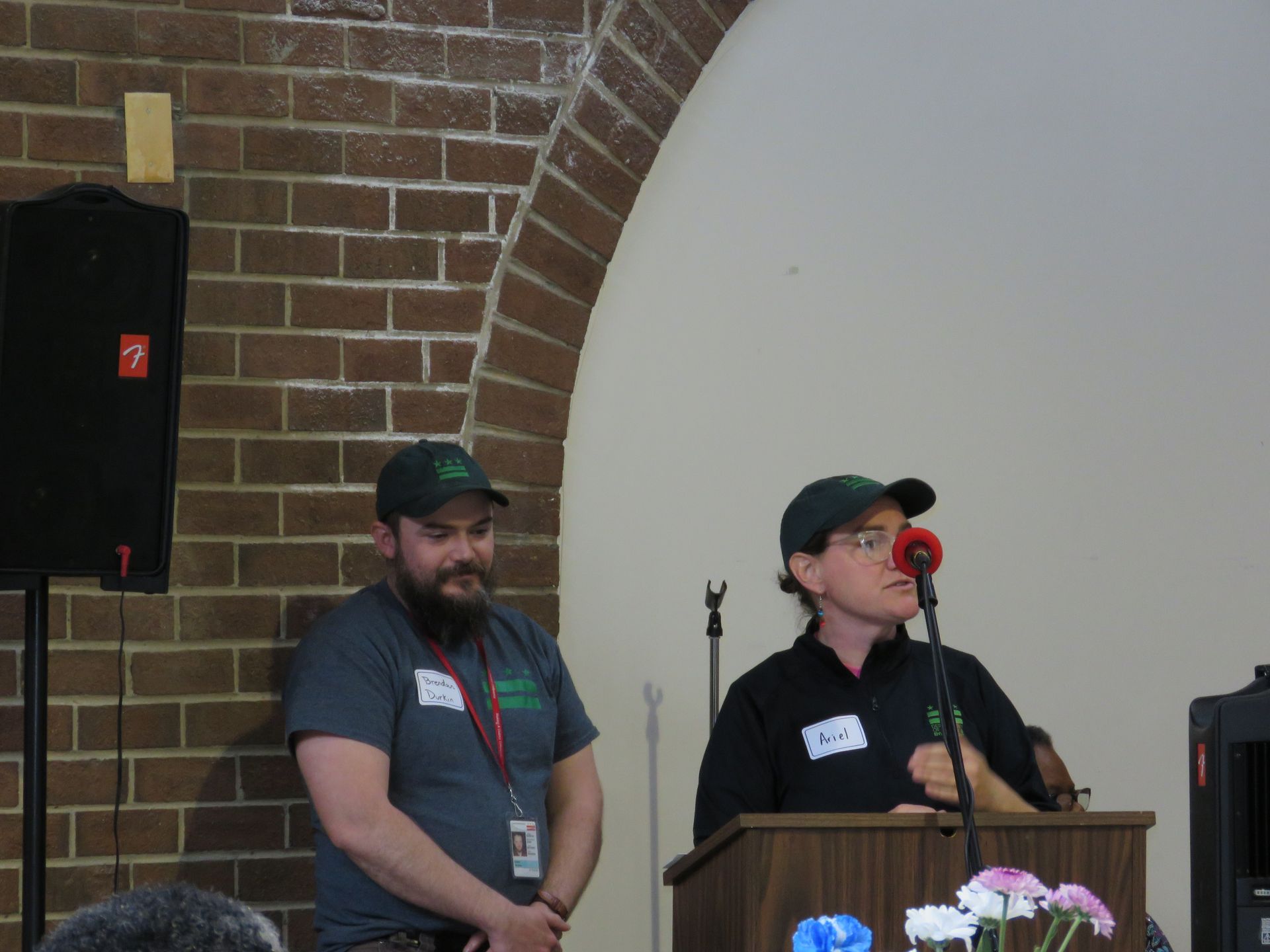 Two people in green hats speak at a podium in front of a brick archway.