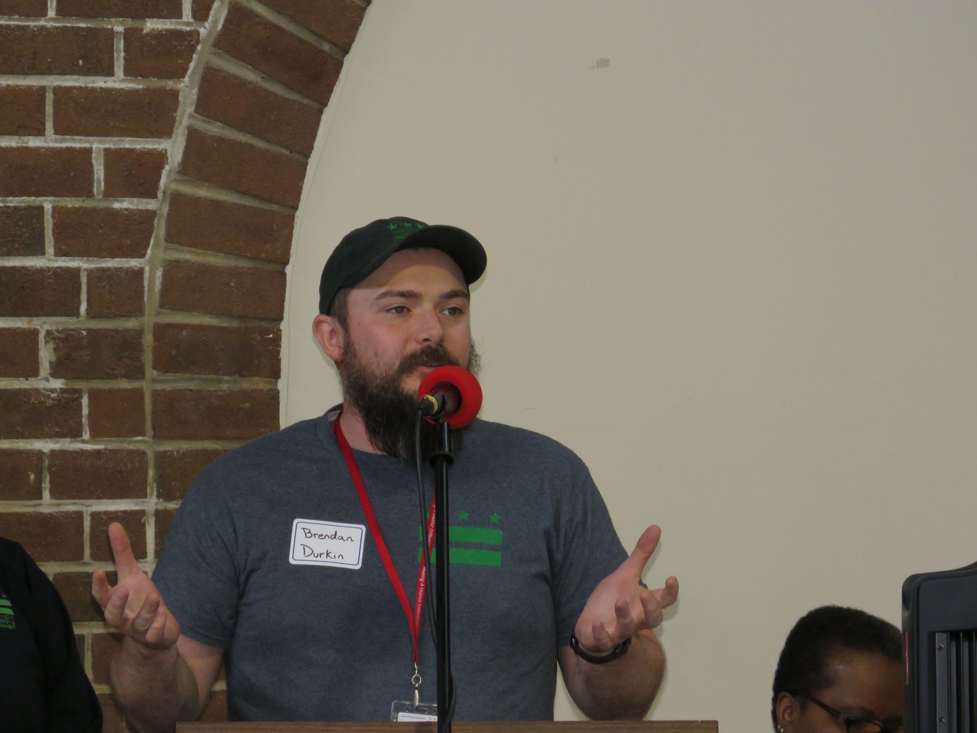 Man with beard speaks at a podium, gesturing with hands. He wears a hat and gray t-shirt. Brick wall backdrop.