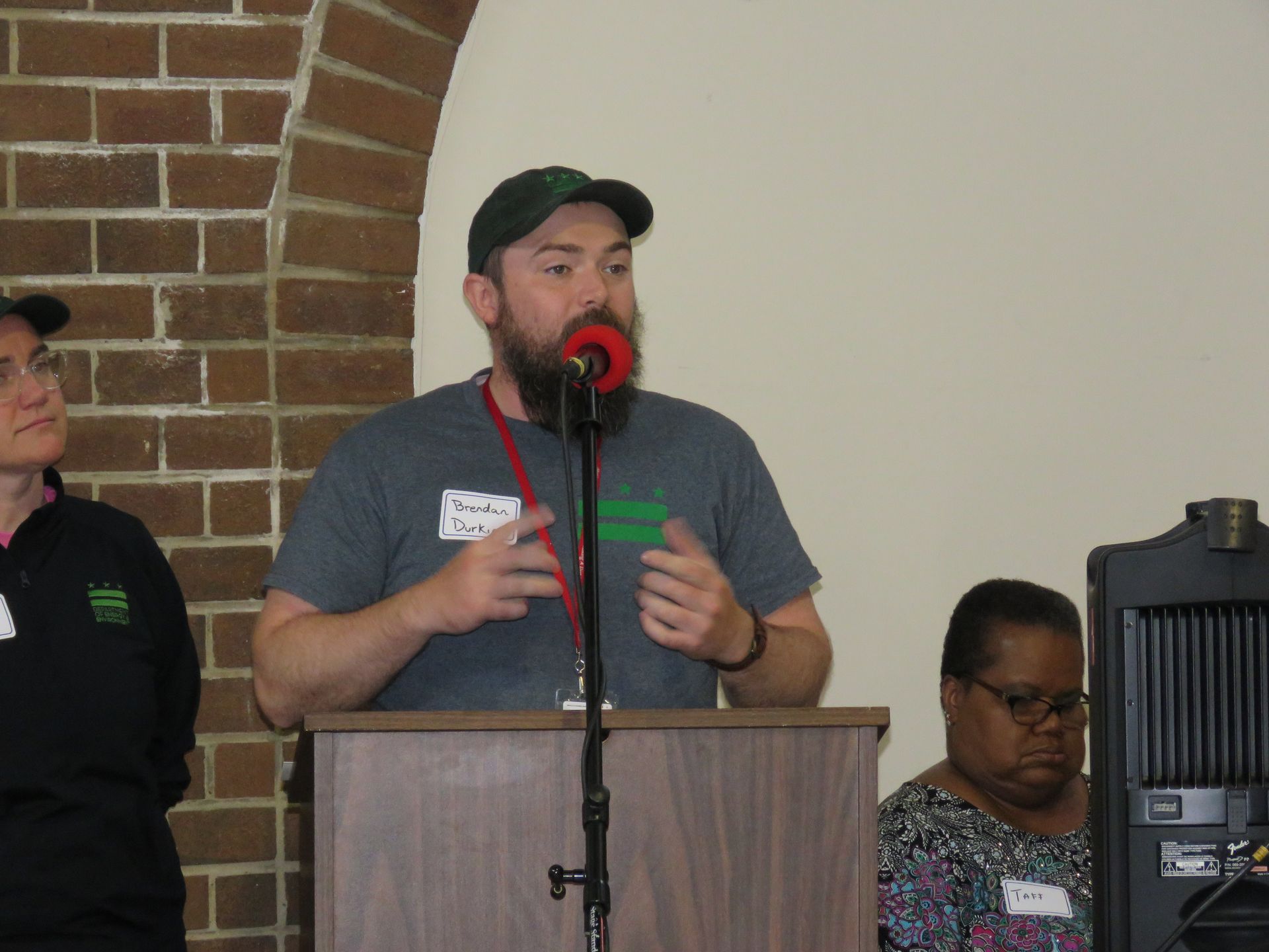 Man with beard speaks at a podium, gesturing with hands; brick archway in background.