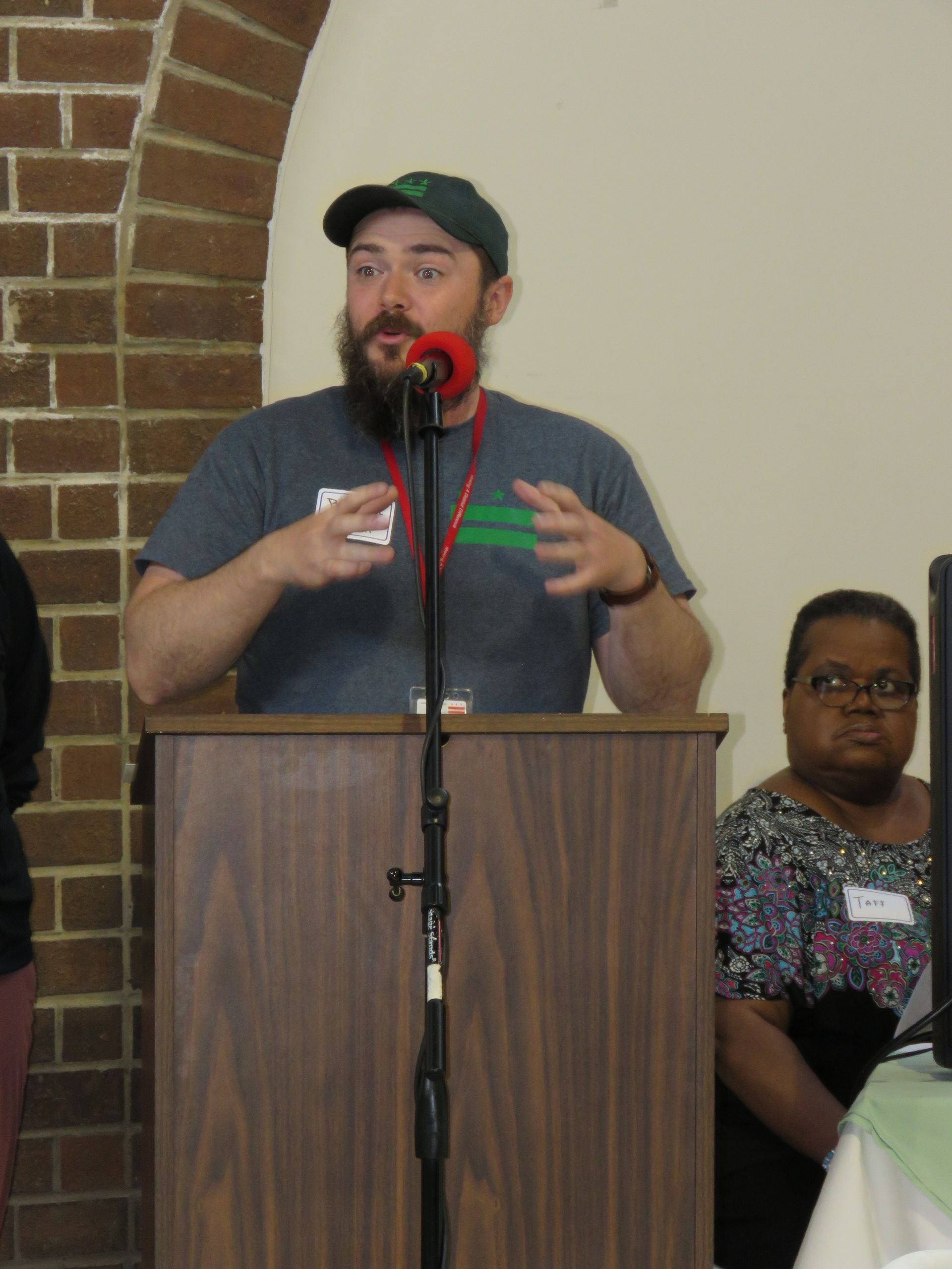Man speaking at a podium, gesturing with hands. He wears a hat and gray shirt. Woman beside him.