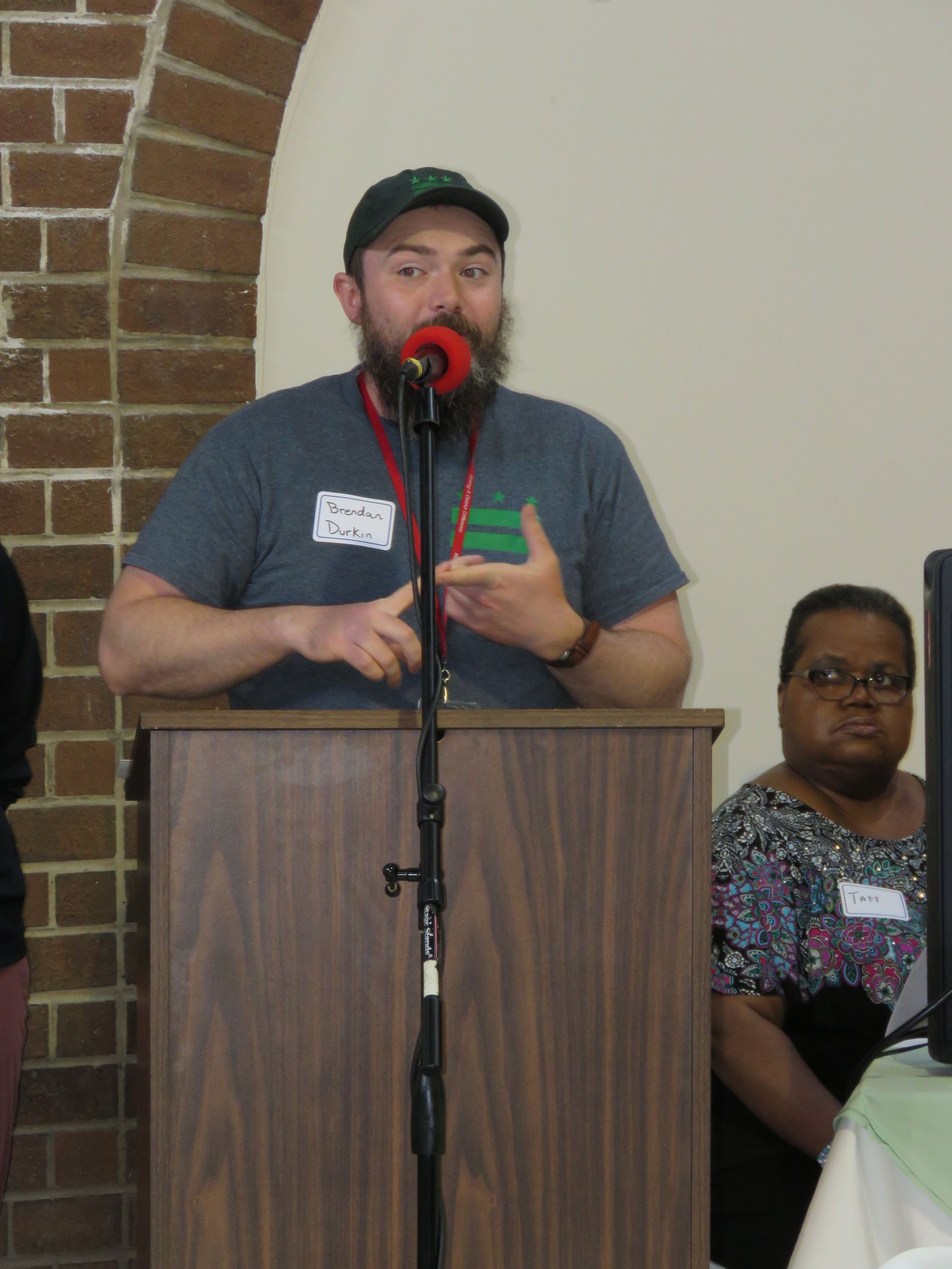 Man with beard speaking at a podium; woman seated nearby, brick wall in background.