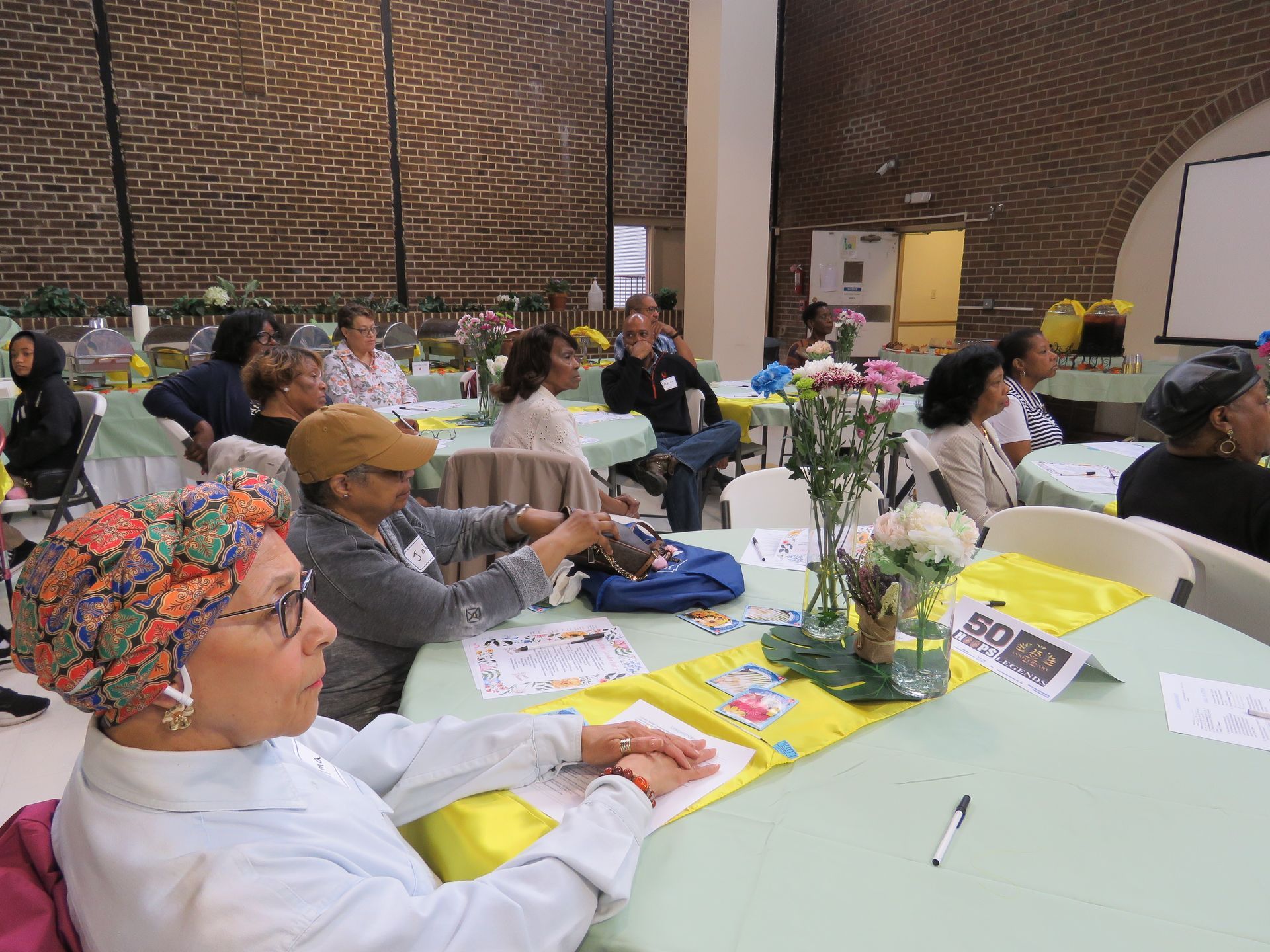 People seated at tables in a room with a presentation, floral decorations, and a brick wall.