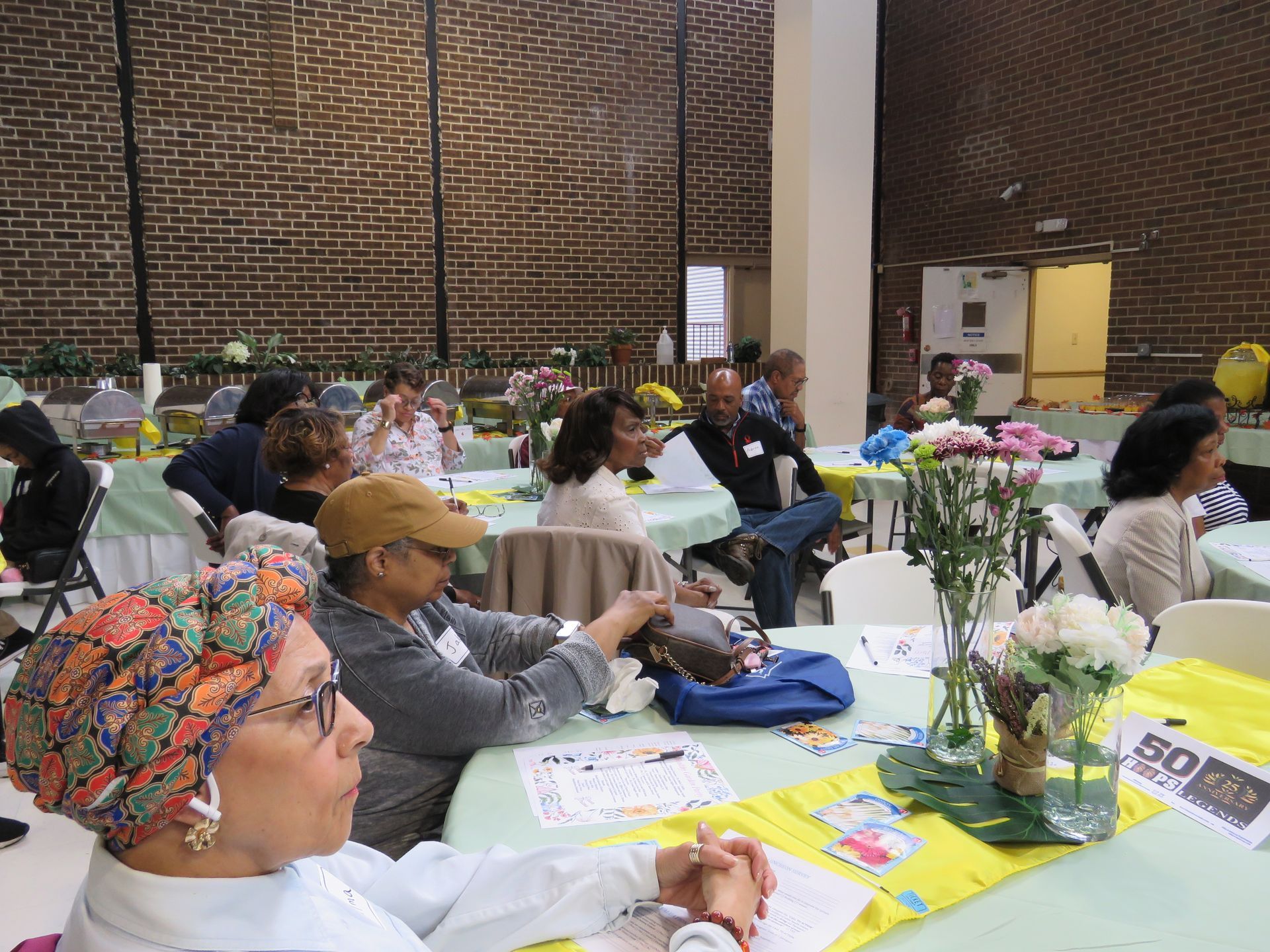 People seated at tables in a room with a brick wall, possibly an event or meeting.