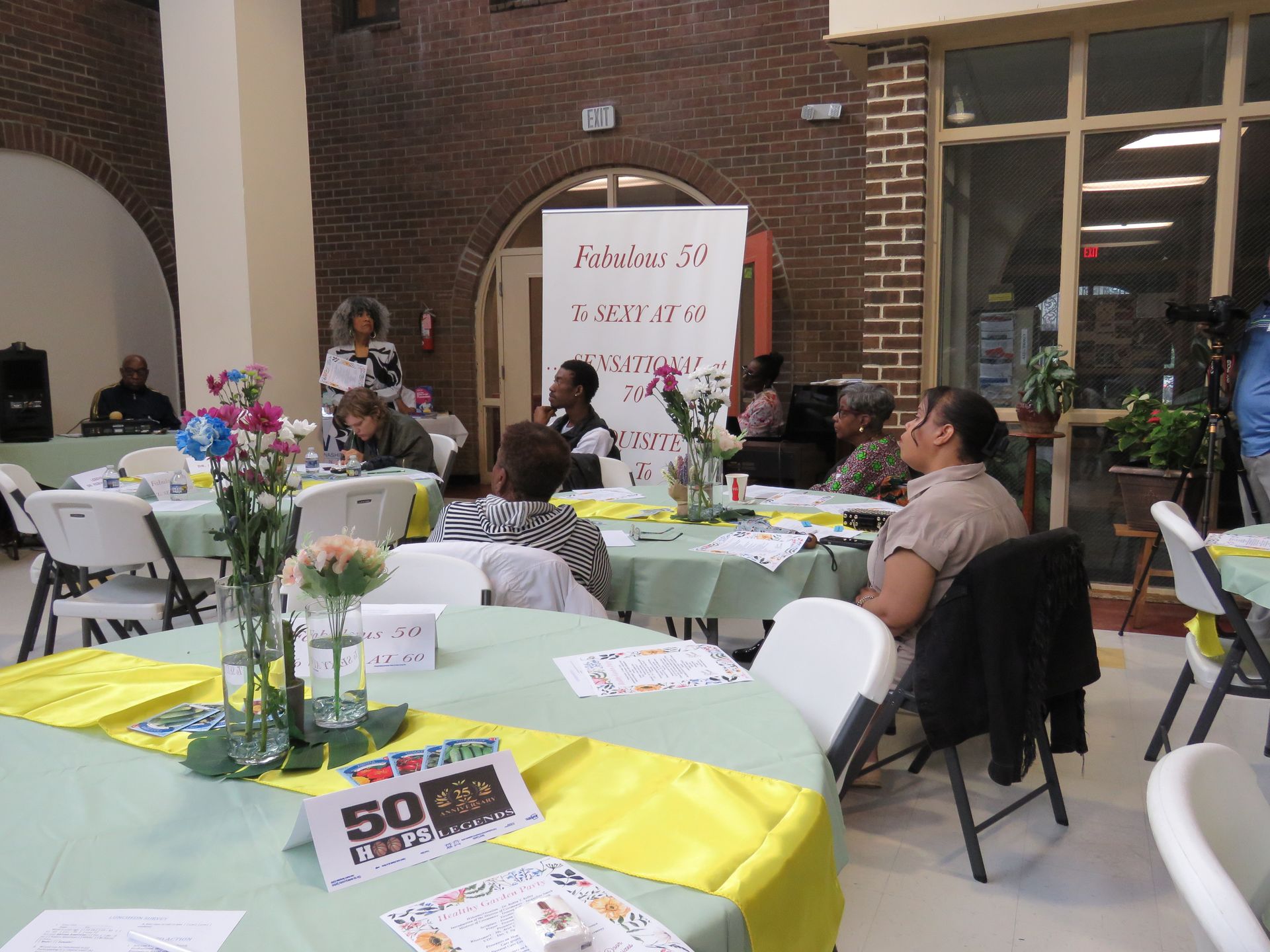 Event attendees seated at tables with decorations, banner, and speaker in a brick-walled room.