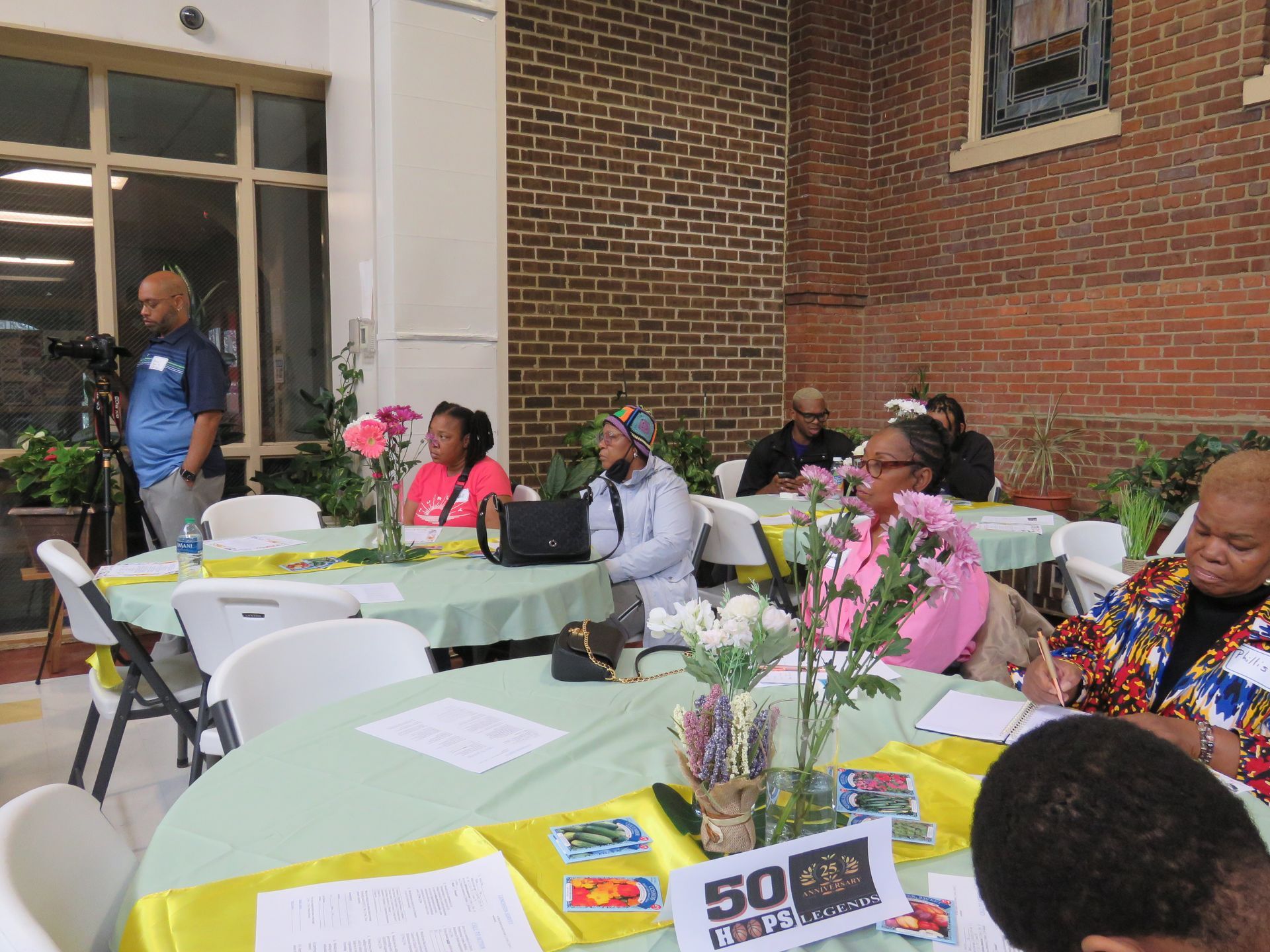 People seated at tables with decorations, listening to a man speaking. Outdoor event with a brick wall.