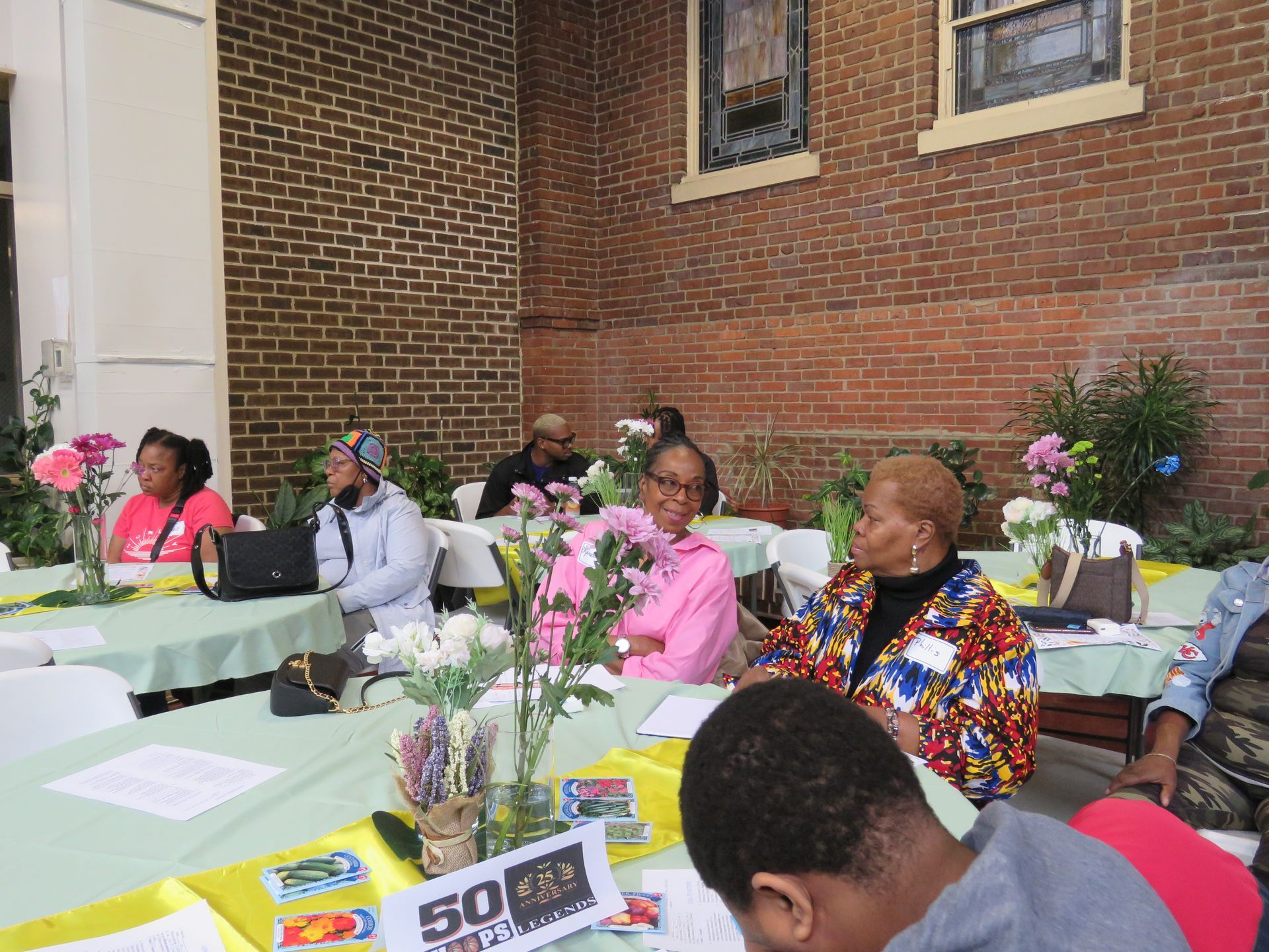 People seated at tables decorated with flowers, in front of a brick building.