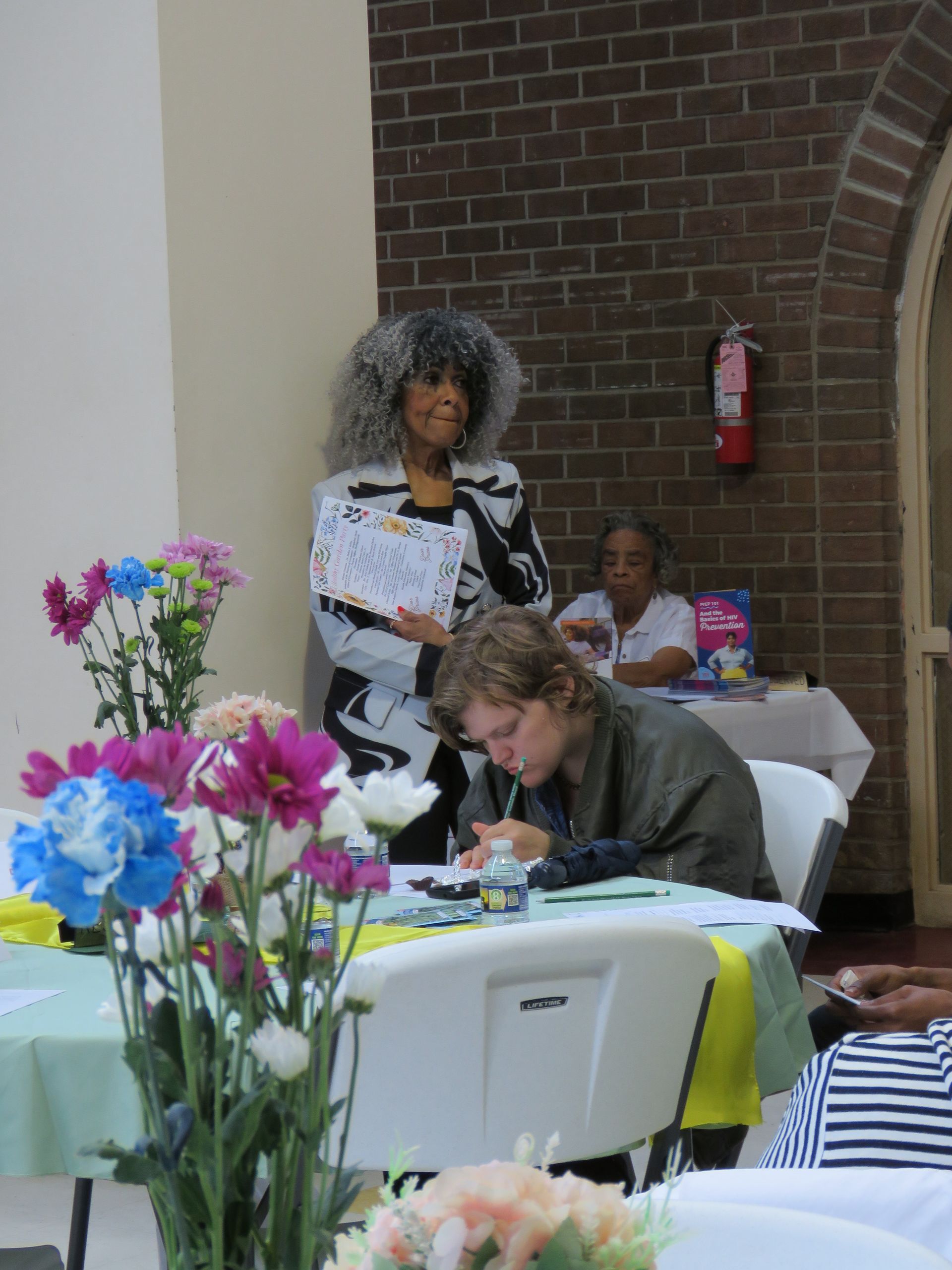 Woman with curly gray hair holds a paper, stands near a person writing at a table with flowers.