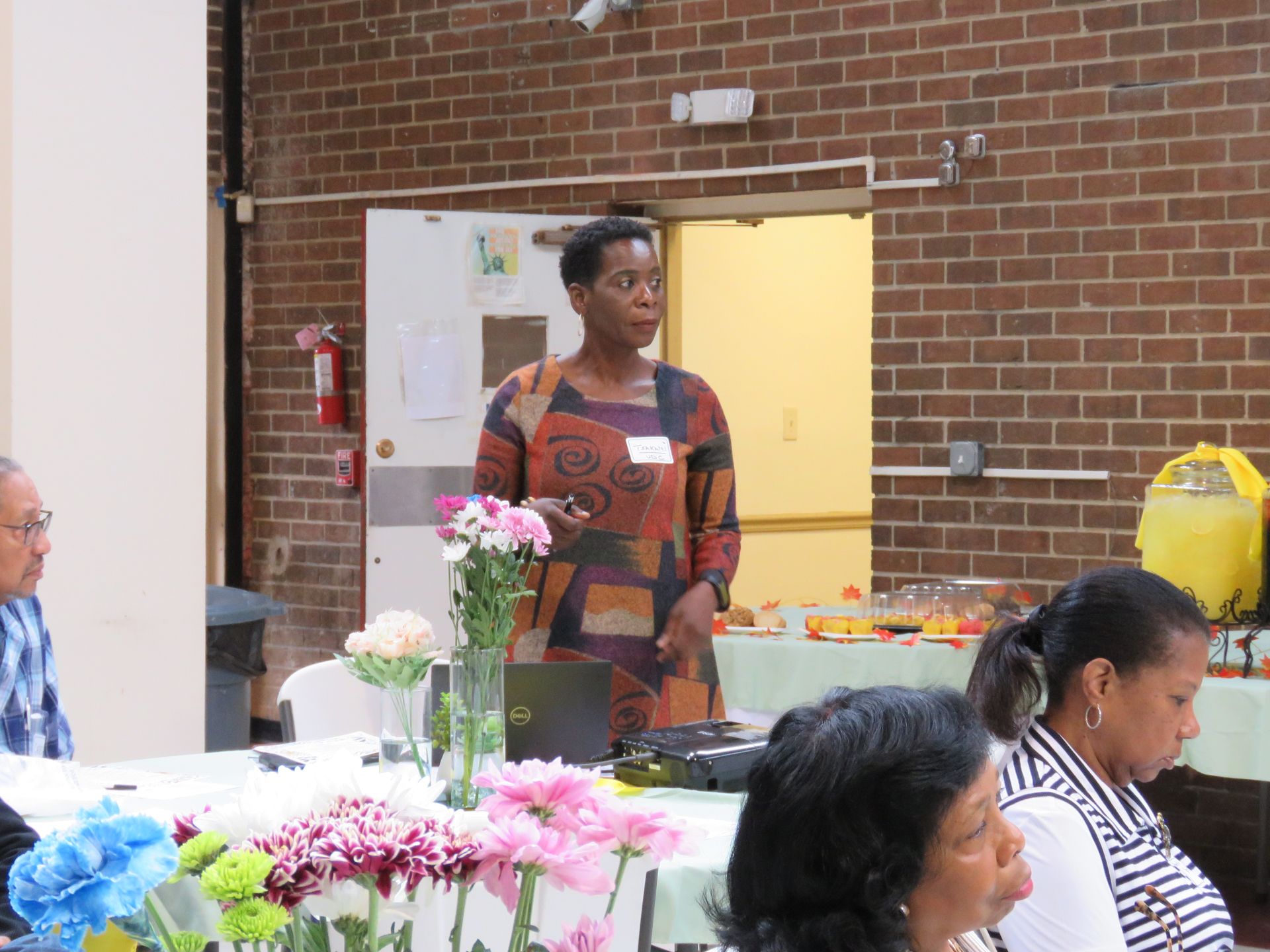Woman giving a presentation at a table, flowers in front, audience present, room with brick walls.