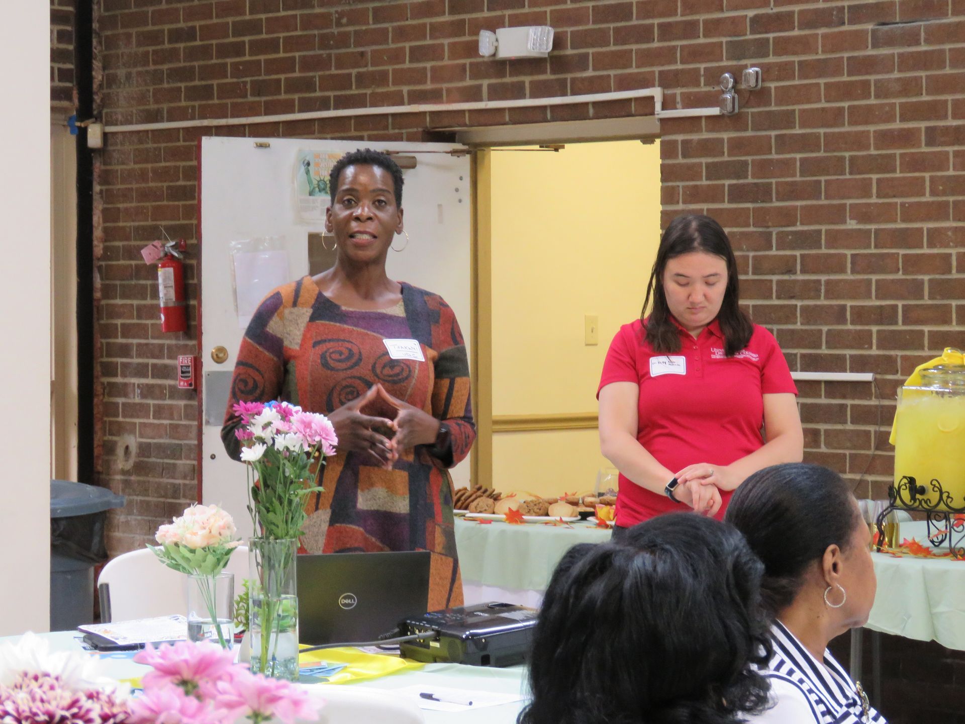 Woman speaking at a presentation, another woman listening. A table with flowers and refreshments is in the foreground.