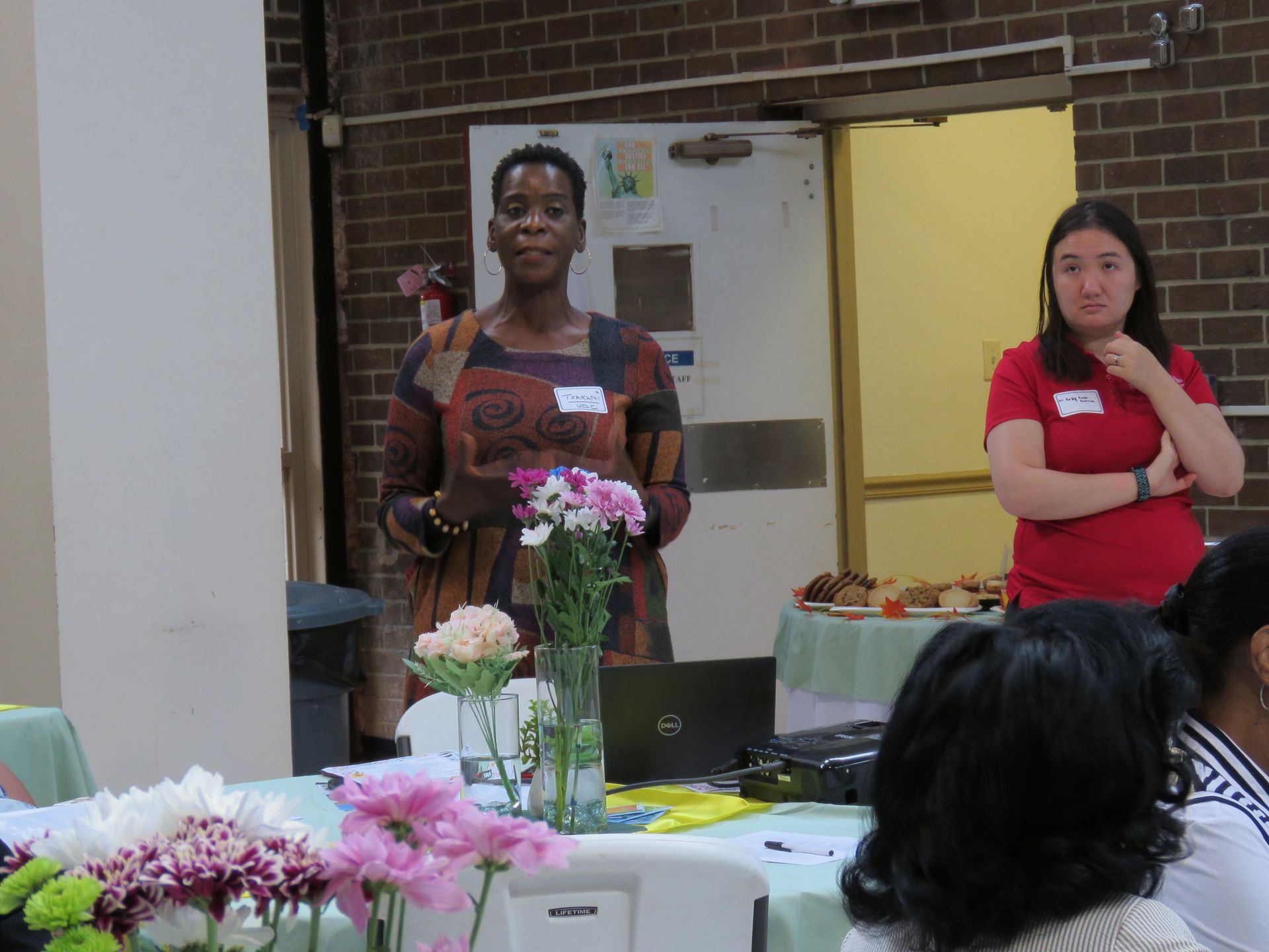 Woman in patterned dress speaking at event. Another woman in red shirt stands nearby. Table with flowers.
